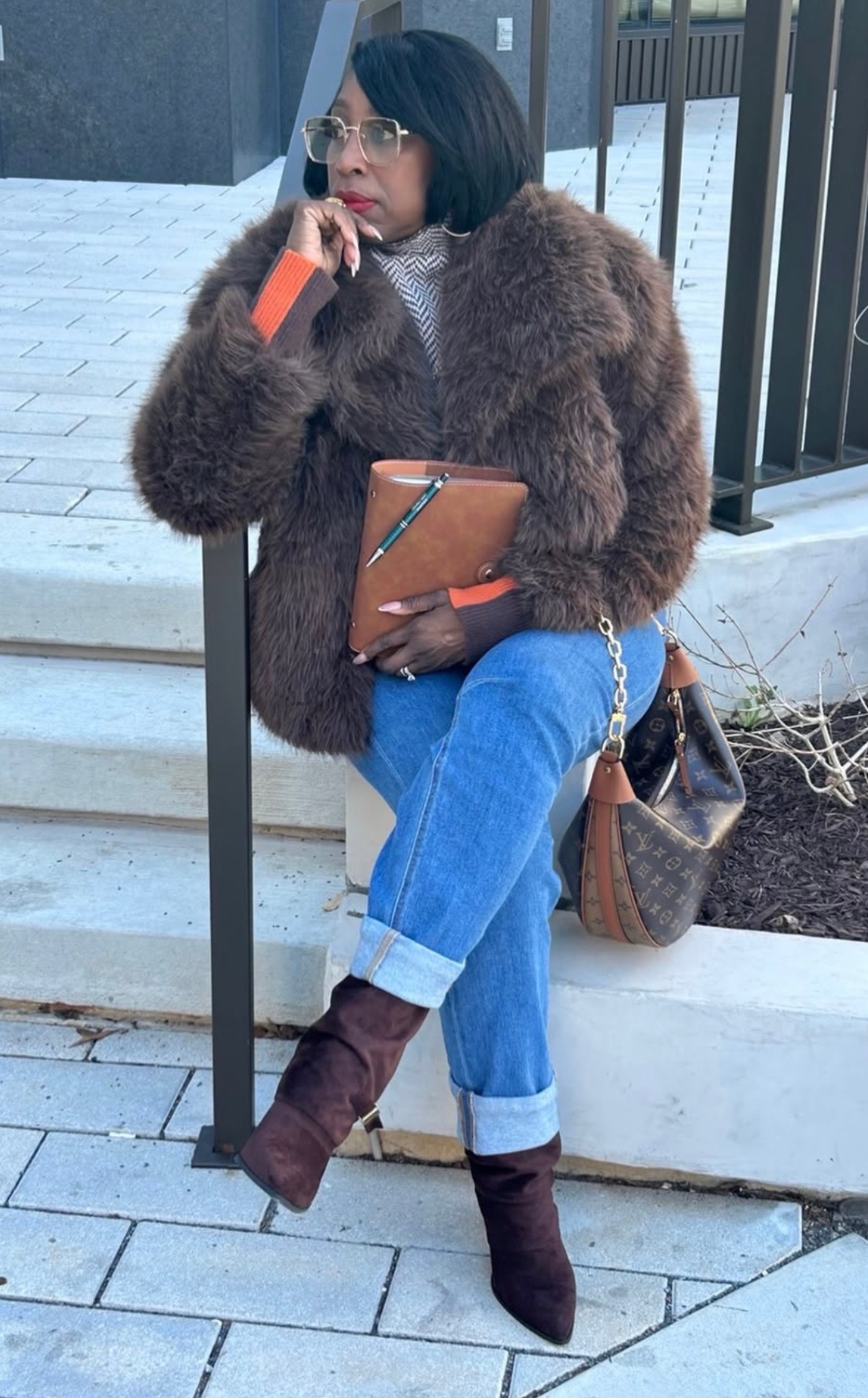 A black woman (Arletta The Friendly Trader) with black bob hair, wearing glasses, a fur coat, and jeans, sitting on a curb with her legs crossed at the ankle, holding a brown leather notebook and a pen, with a Louis Vuitton handbag beside her.