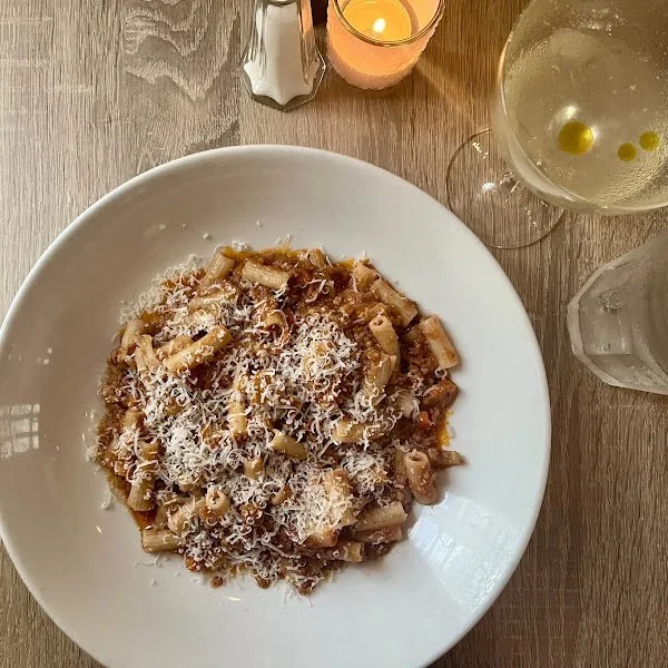 A plate of pasta from Pasta da Pulcinella with grated cheese, a glass of white wine, a glass of water, a lit candle, and a salt shaker on a wooden table.