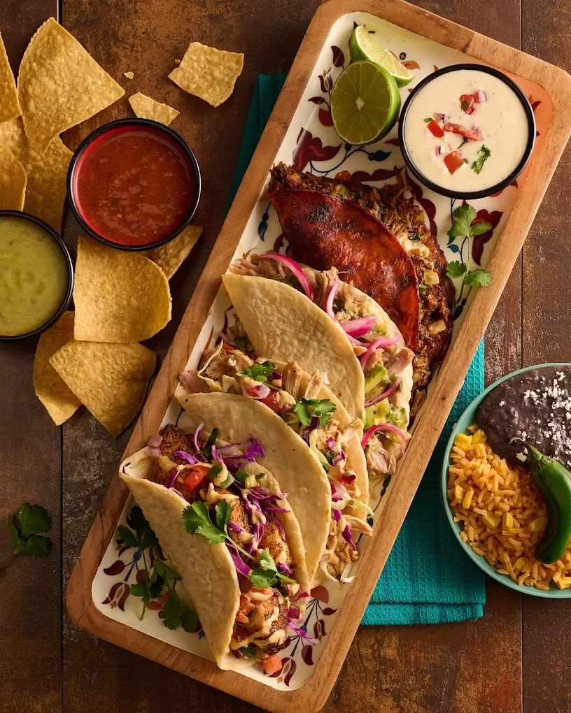 A wooden platter from Dos Caminos with three tacos filled with shredded meat and vegetables, a grilled jalapeño, a lime wedge, and a small bowl of creamy sauce topped with chopped peppers.