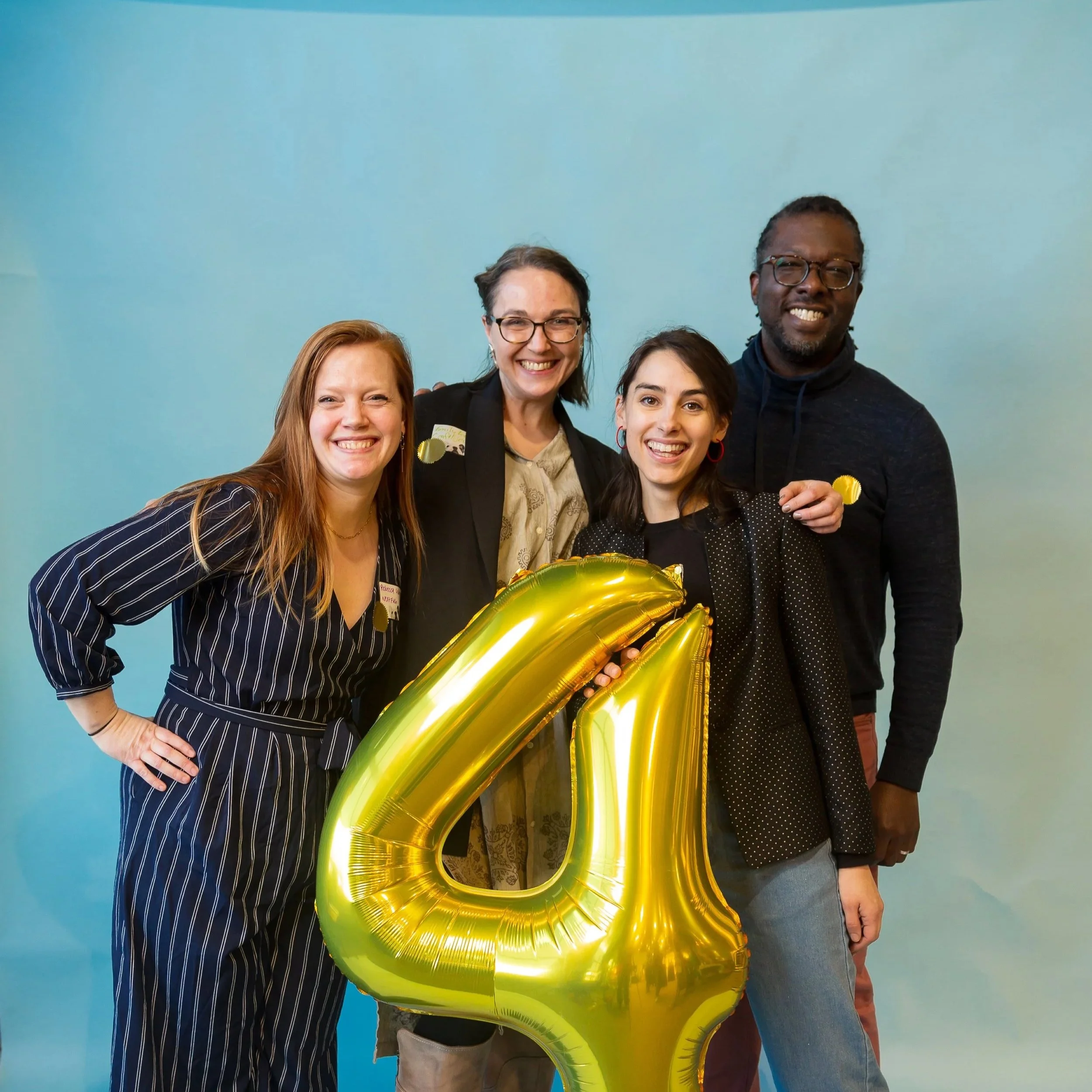 Four people smiling and celebrating a 4th anniversary, holding a large gold balloon shaped as the number 4, against a light blue background.