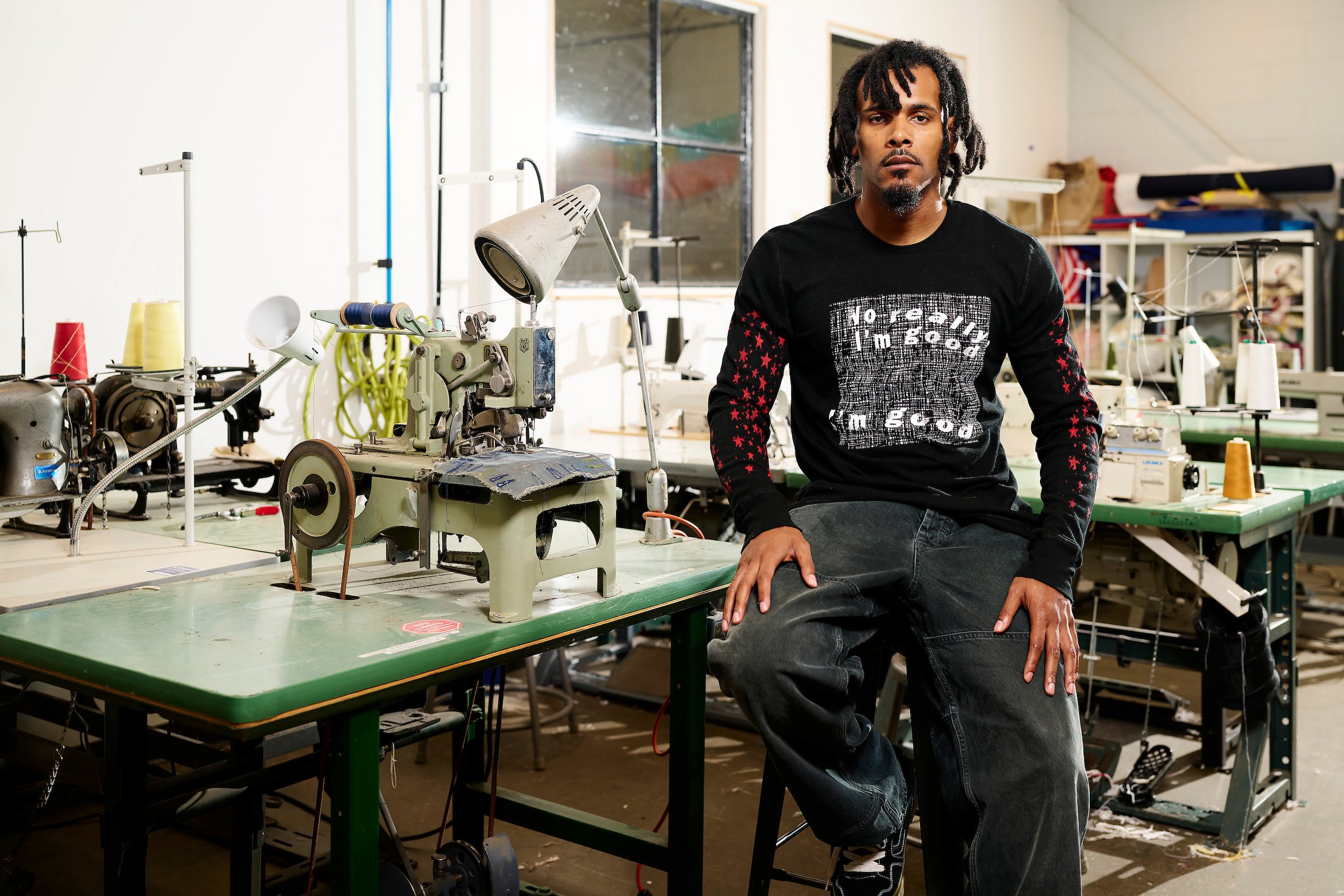 A man with dreadlocks sitting on a stool in a workshop with sewing machines and fabric on tables and shelves.