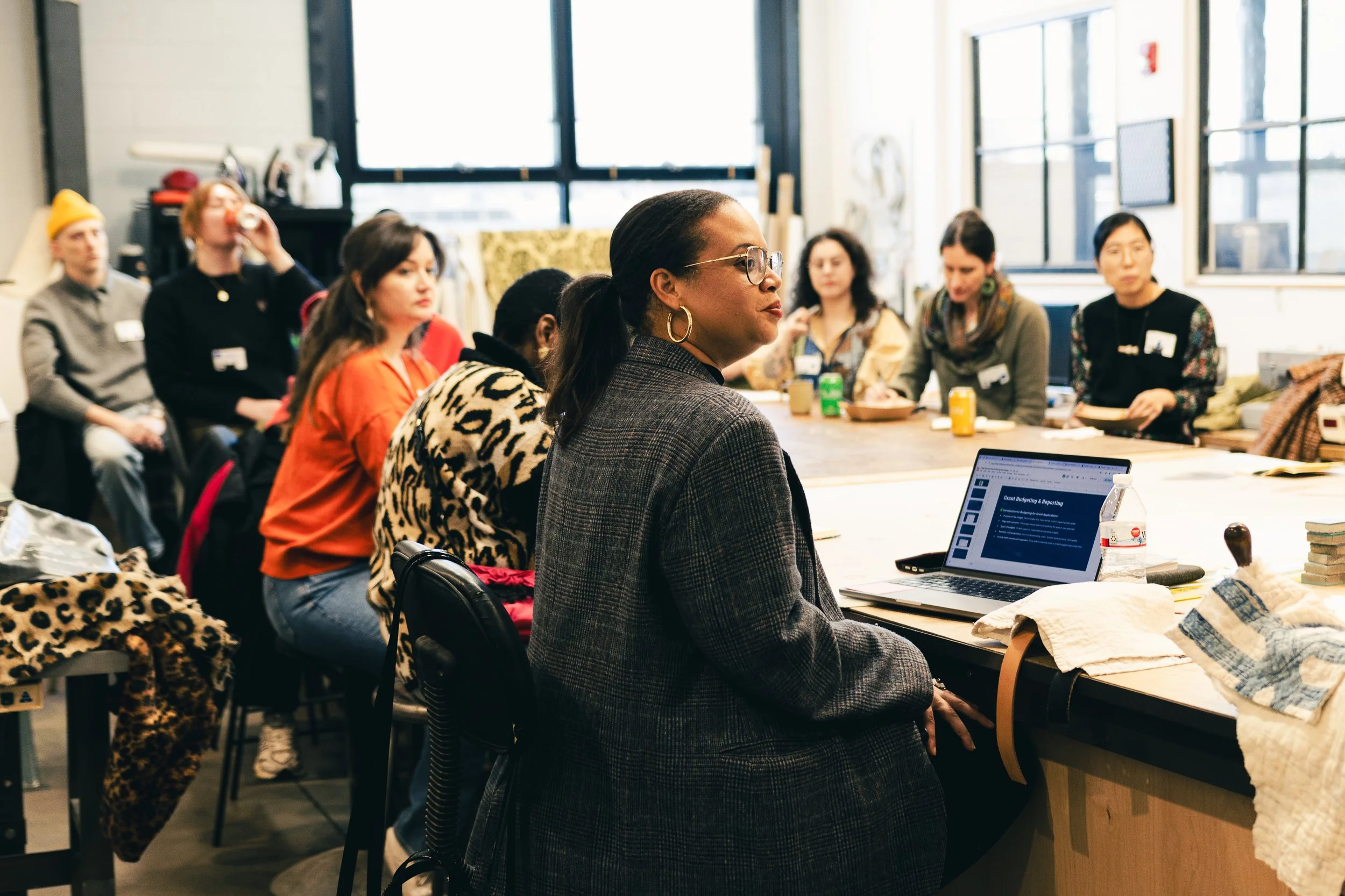 A group of diverse women gathered around a table in a bright workshop or classroom, listening to a woman at the front who is wearing glasses and a plaid blazer, with a laptop open in front of her.