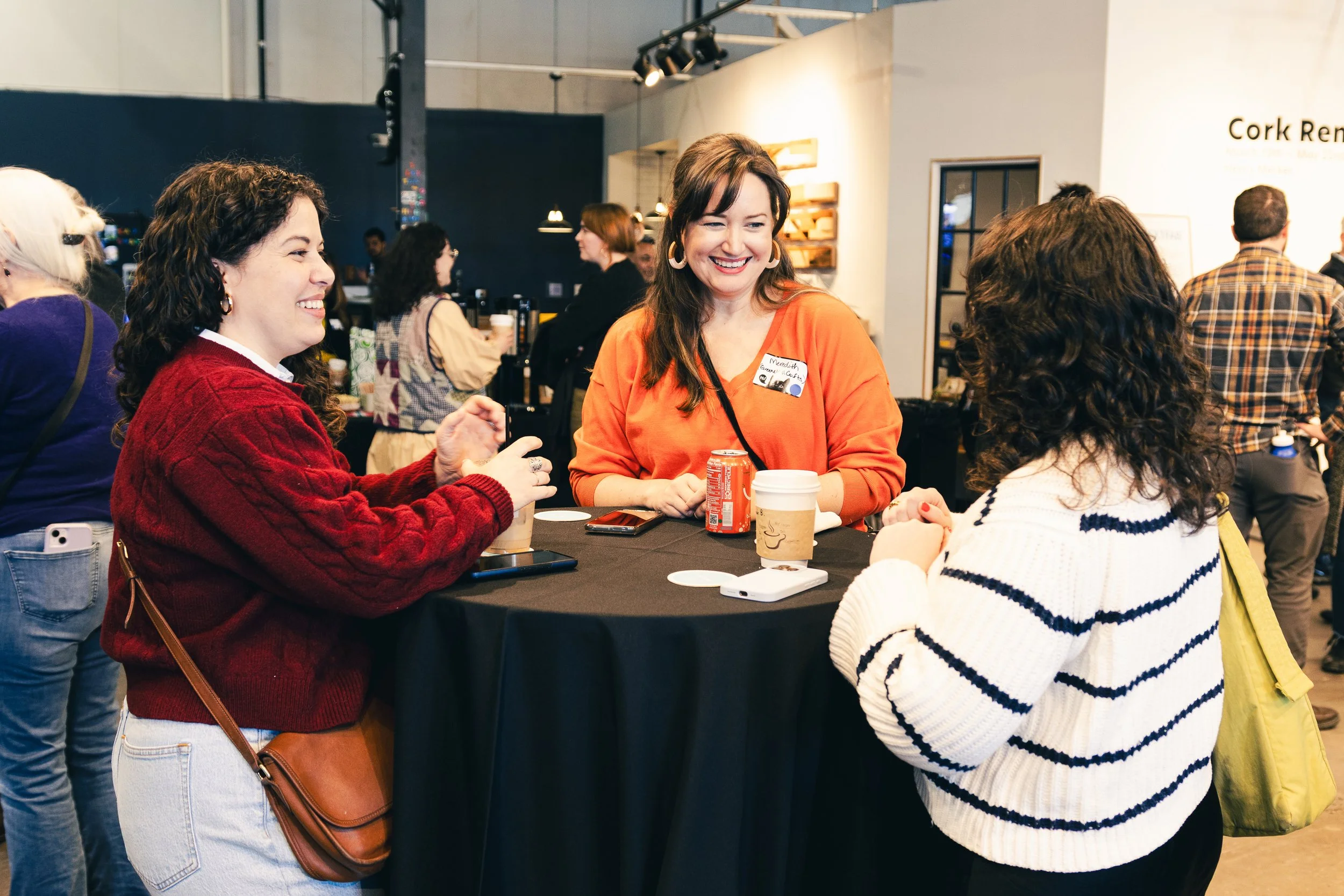 Three women smiling and chatting around a high table at an indoor event, with coffee cups and a soda can on the table, and a crowded background with other people.