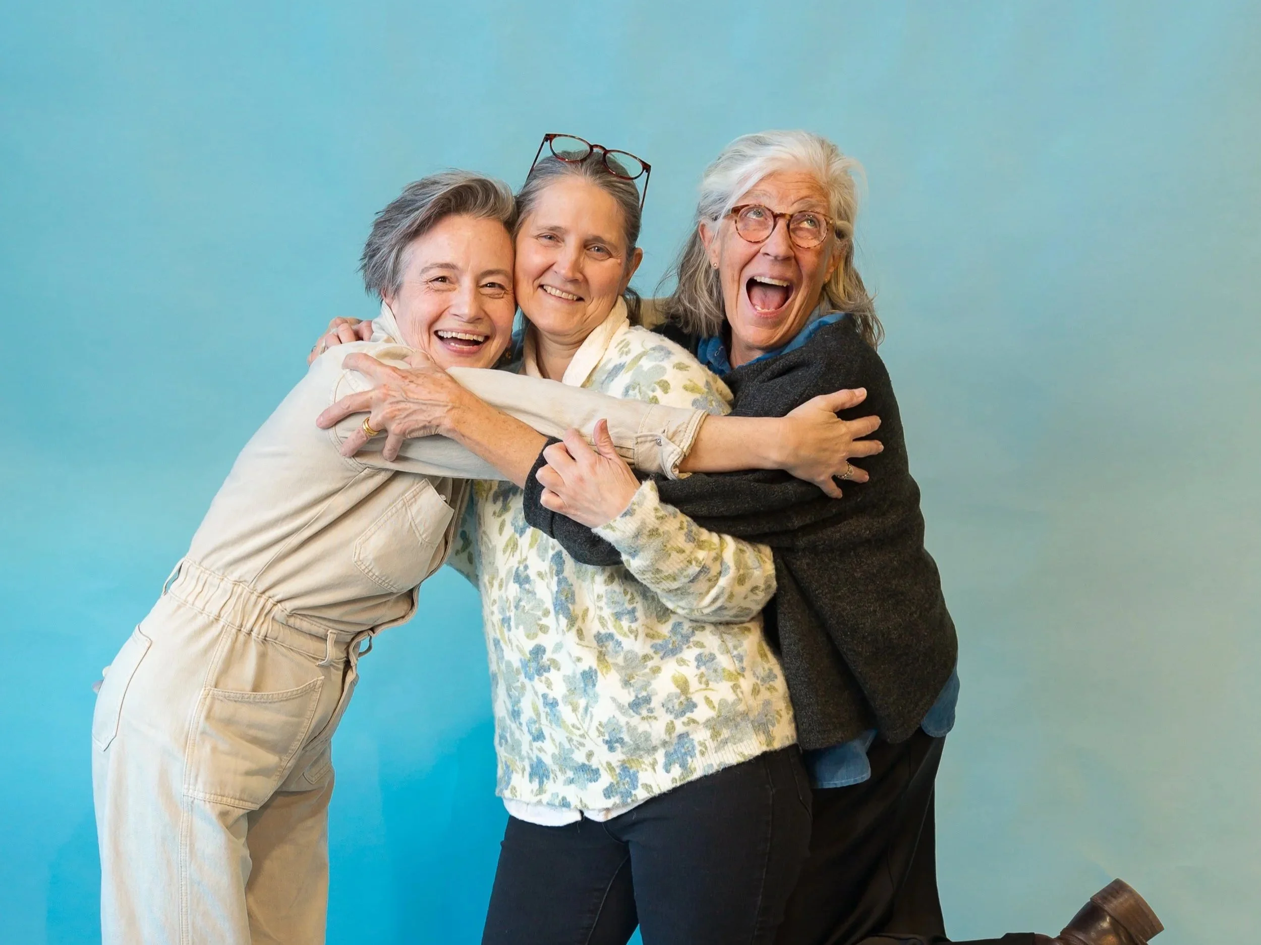 Three women hugging and smiling against a light blue background.