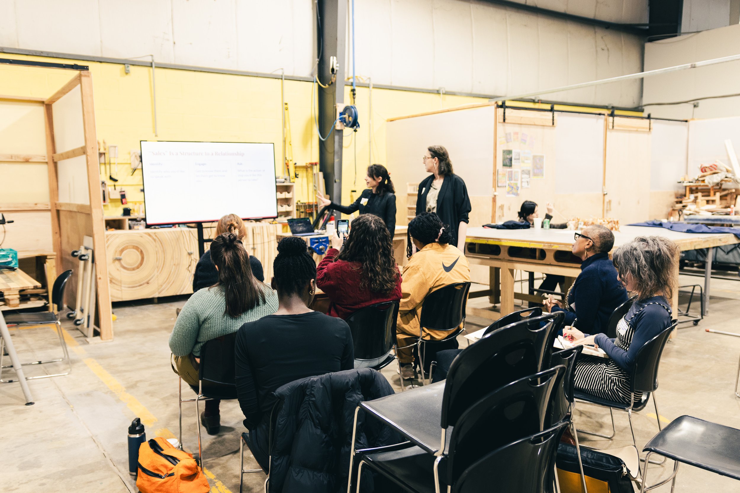 A group of people attending a workshop or presentation in a workshop space with wooden tables and machinery, watching two women at the front giving a presentation on a large screen.