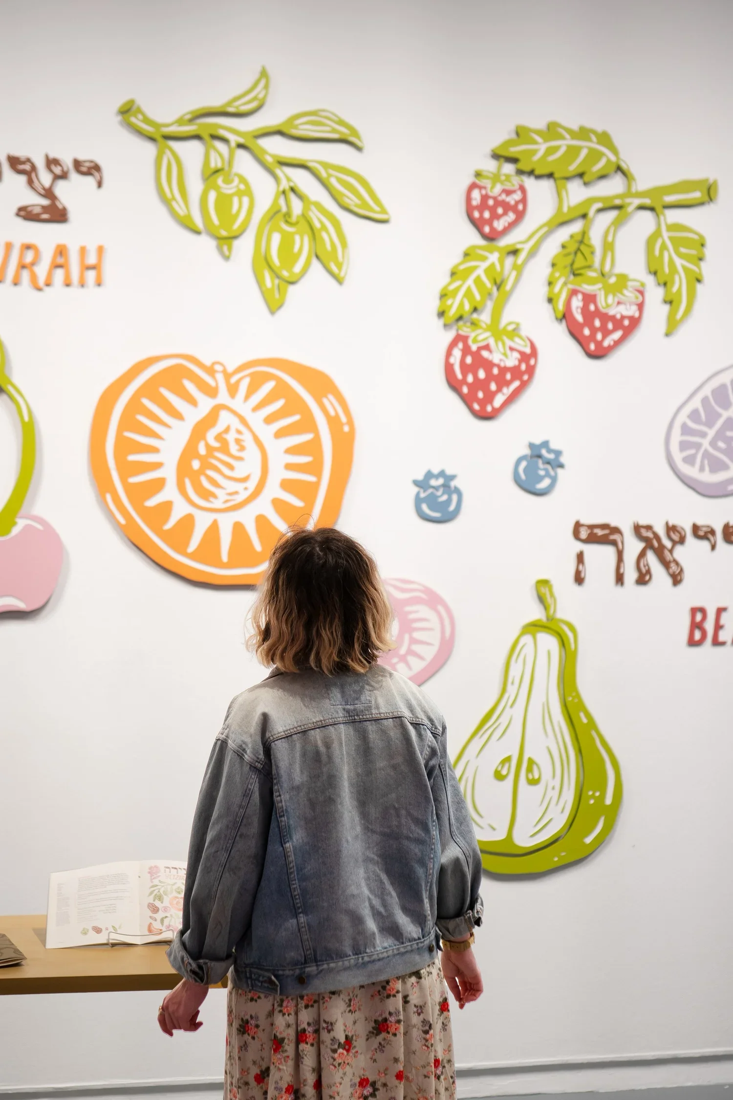 A woman stands in front of a colorful wall decoration of various fruit illustrations, including strawberries, oranges, pears, and blueberries, with some text in a different language.