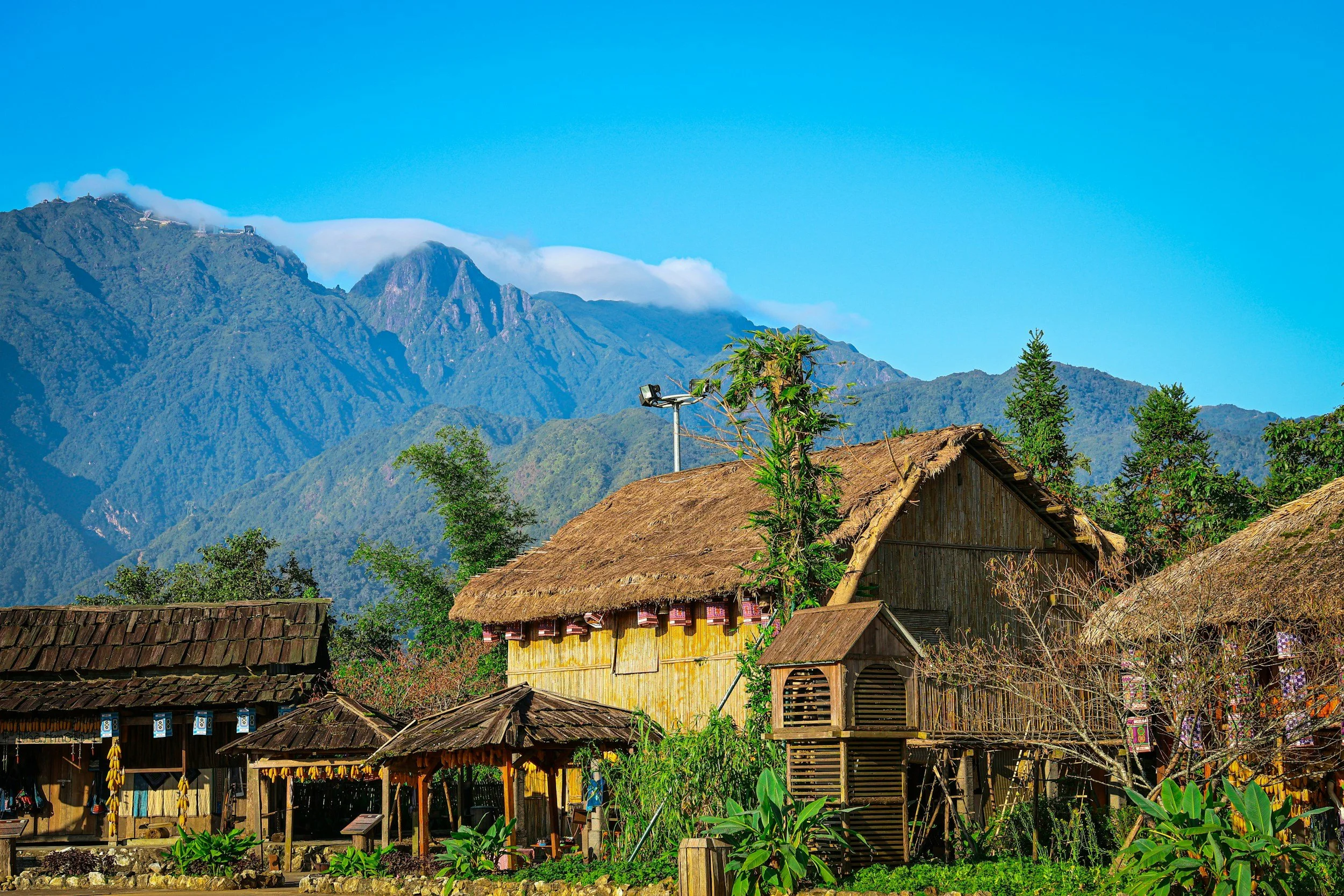 A rural village with bamboo houses and surrounding greenery, set against a backdrop of lush mountains and a clear blue sky.