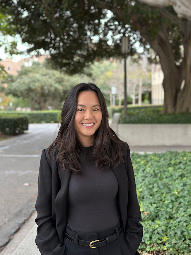 Smiling woman with long dark hair, wearing a black blazer and black top, standing outdoors in a park.