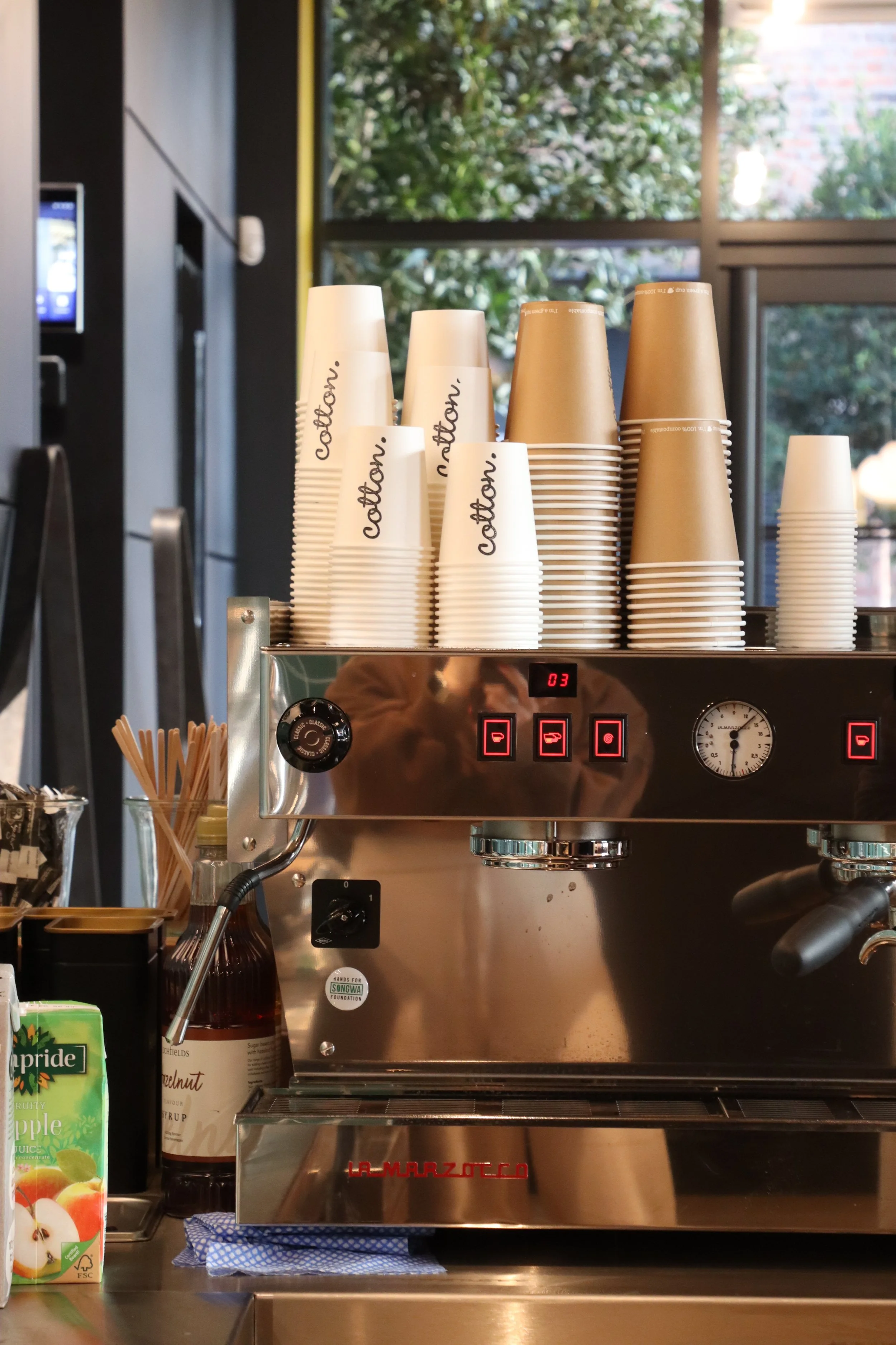 A commercial espresso machine with stacked paper cups on top, some labeled 'caffeine.' Bottles, straws, and juice boxes are on the counter in front of it in a cafe setting, with windows showing greenery outside.