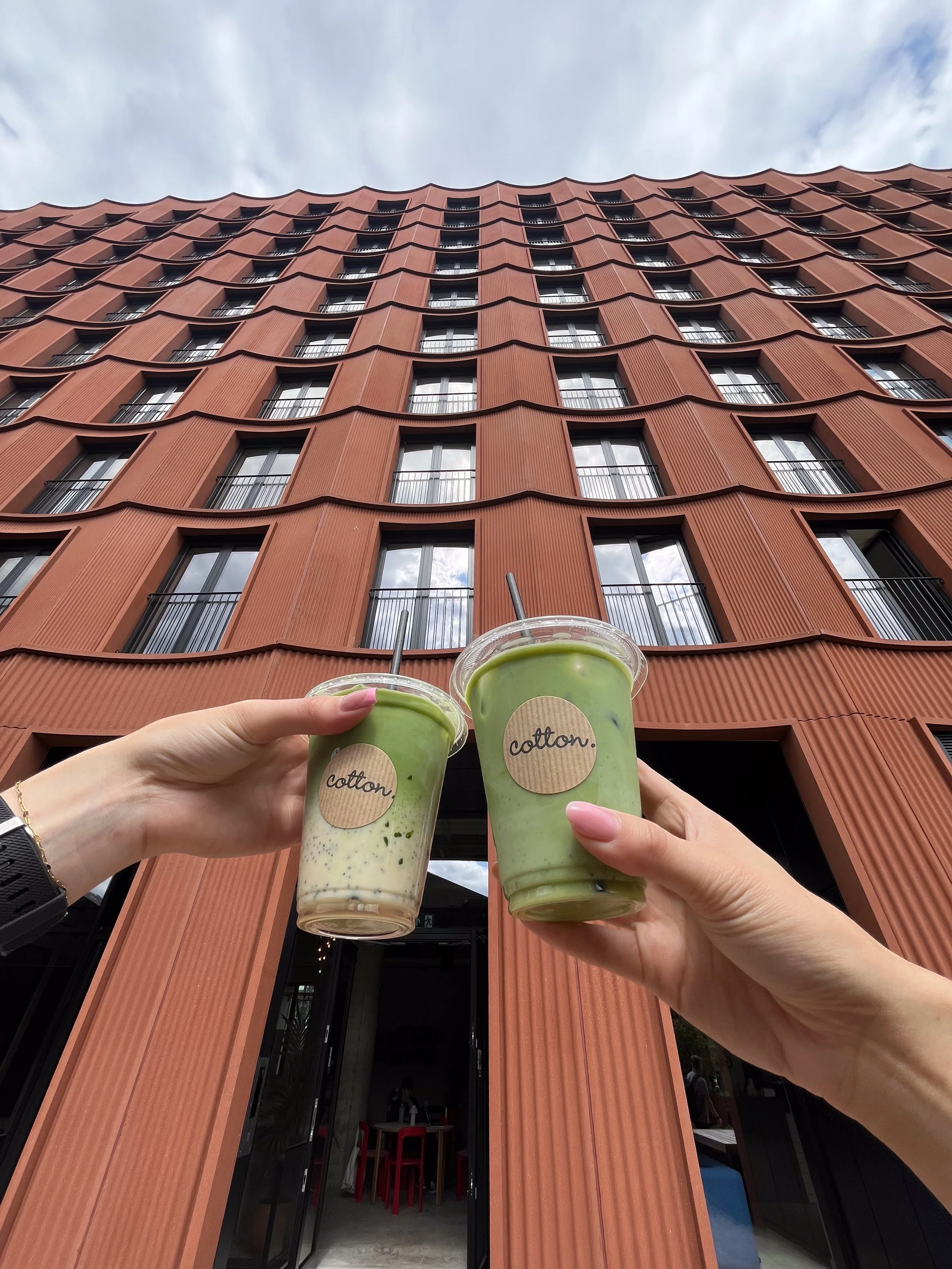 Two people holding cups of green beverages outside a modern, multi-story building with a wavy, orange facade and large windows.