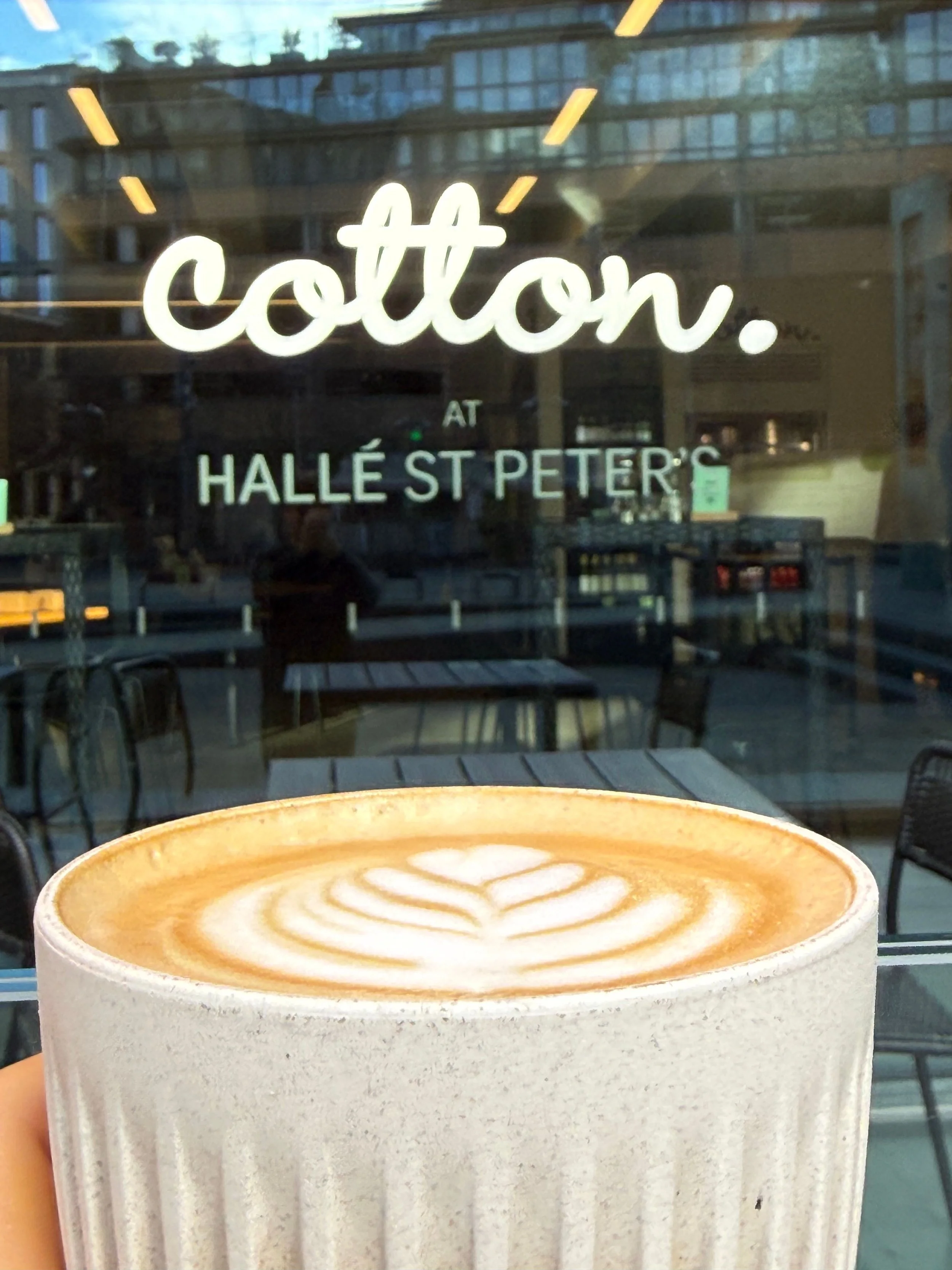 A cup of latte with latte art in front of a glass window with neon sign reading 'cotton. at Hallé St Peters'.