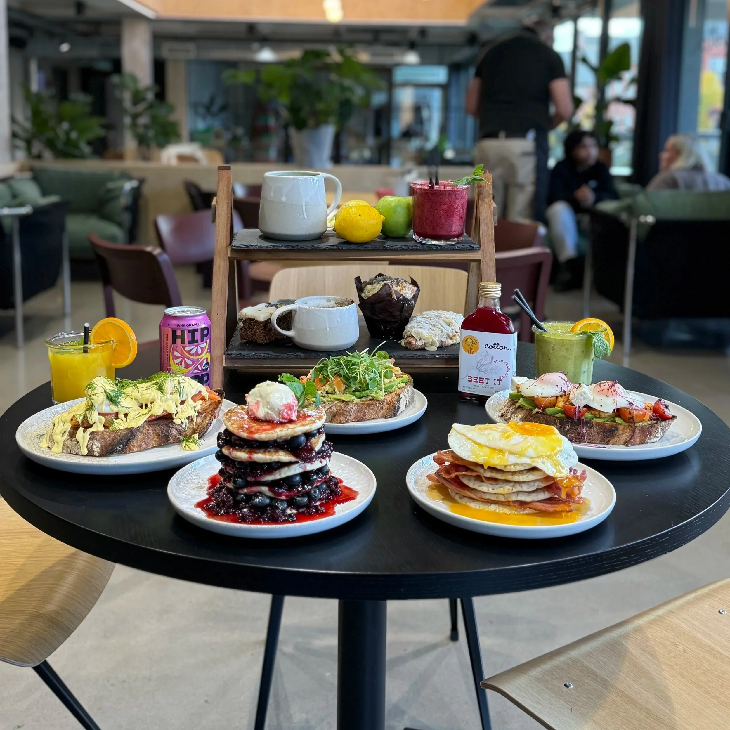 A table in a cafe set with various breakfast foods including pancakes with berries, avocado toast, eggs with ham, and toast with salmon and poached eggs, accompanied by drinks such as juice, smoothie, and soda; a multi-tiered serving stand with cups, lemons, and a smoothie on top, and jars of jam, with a cozy cafe background.