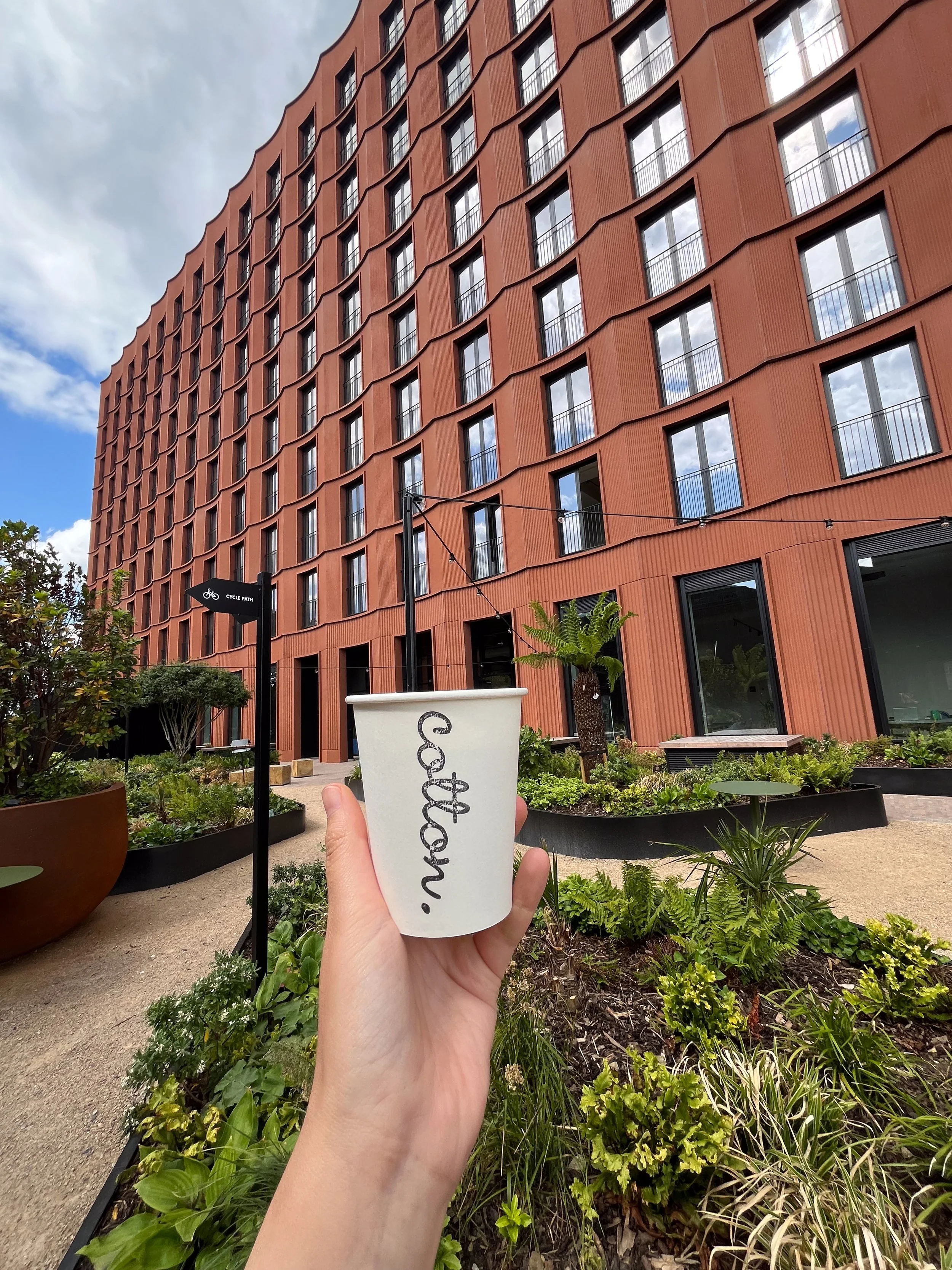 A person holding a white cup with the word ‘Coffee.’ written on it, in front of a modern terracotta-colored building with many windows and balconies, surrounded by landscaped greenery and a paved pathway, under a partly cloudy sky.