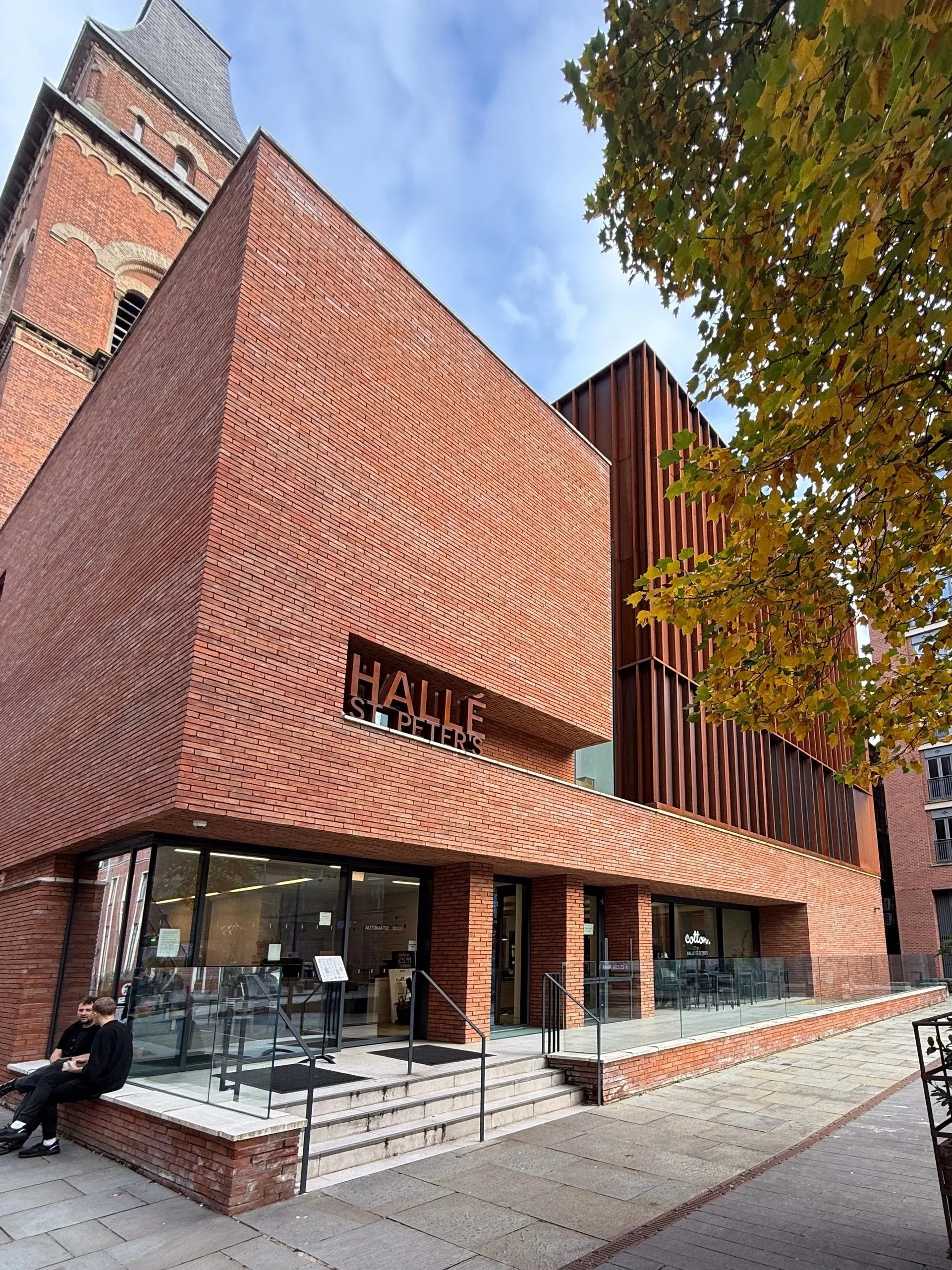 A modern brick building with glass doors and signage for Saint Peter's Hall, located on a city sidewalk with people sitting outside and a tree with autumn leaves.
