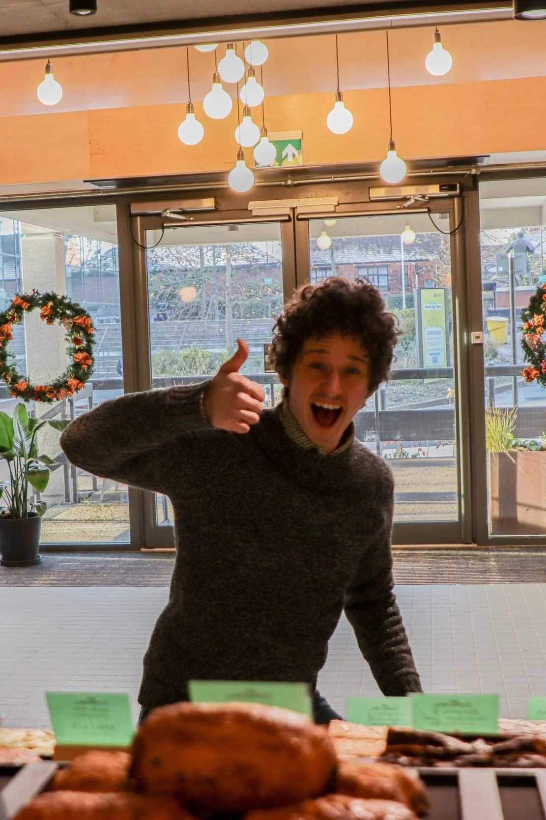 A young man with curly hair giving a thumbs up and smiling inside a store or cafe, with a display of baked goods in front of him. The background features glass doors and windows, Christmas wreaths, and pendant lights.
