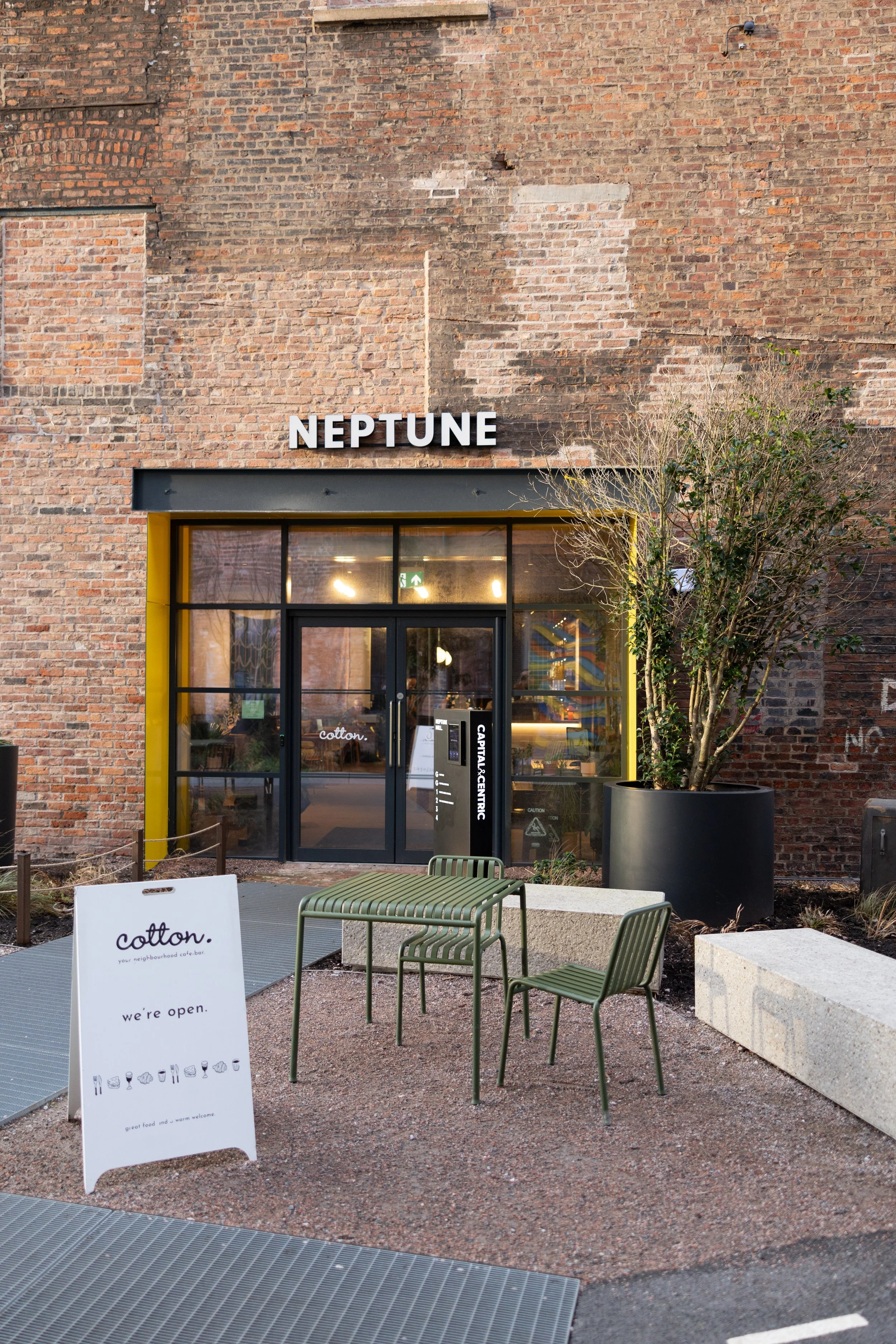 Exterior of a modern cafe called Neptune with a brick wall background, large glass windows, and a black and yellow frame. Outdoor seating includes green striped chairs and a white signboard advertising the cafe's opening.