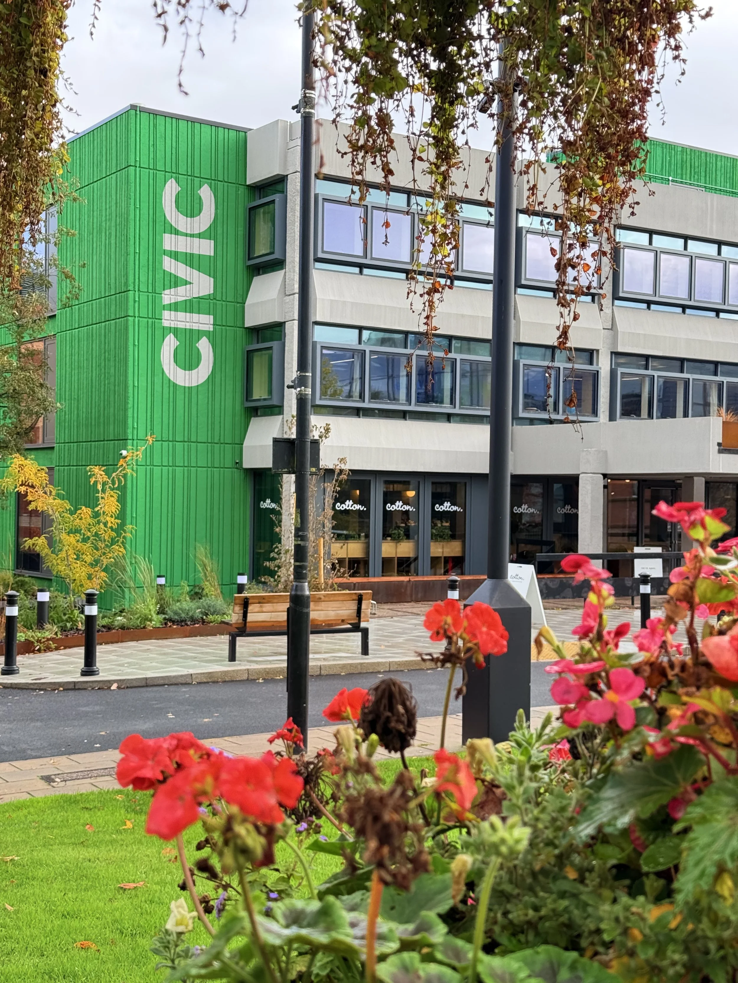 Multistory building with green vertical siding on one corner, large windows, and a sign that reads 'CIVIC'. Outdoor seating area with benches, potted plants, and a well-maintained lawn and flower beds in the foreground.