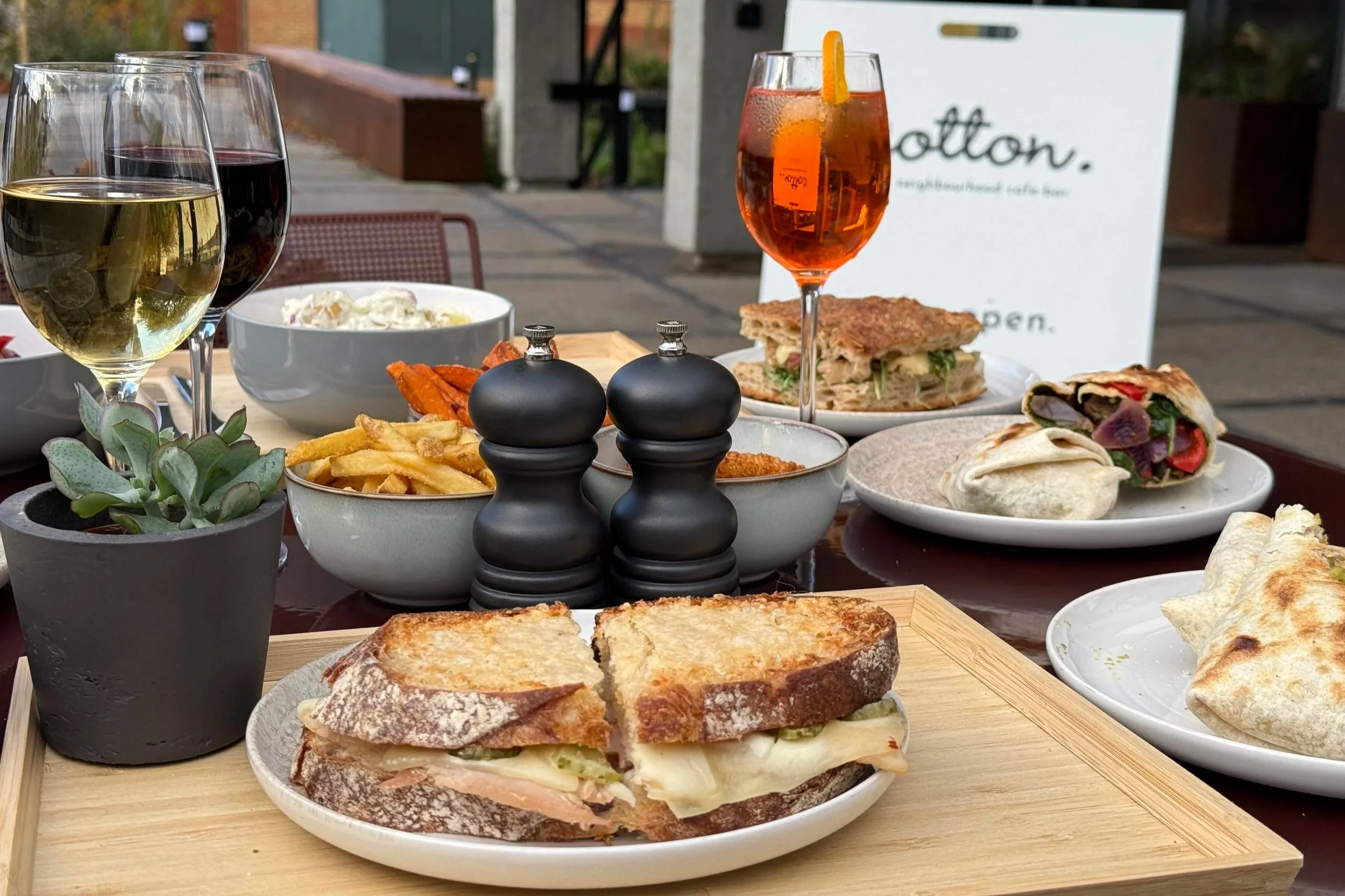 An outdoor restaurant table set with sandwiches, fries, wine glasses, a cocktail, and various side dishes, with a tree and building in the background.