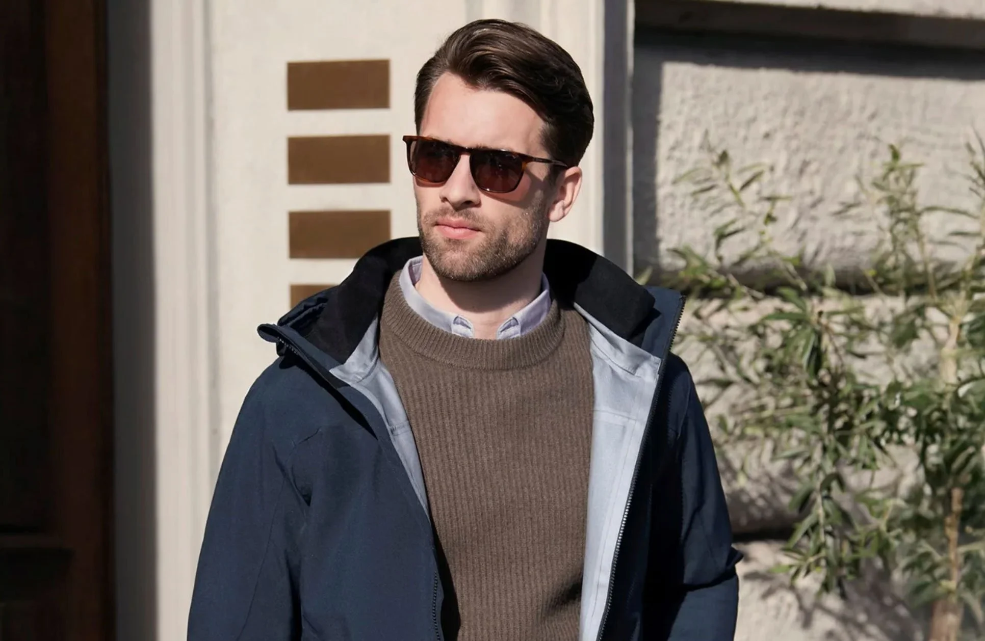 Ung mann med brunt hår, skjegg, solbriller, iført blå jakke, hvit skjorte og brun genser, står utendørs i sollys