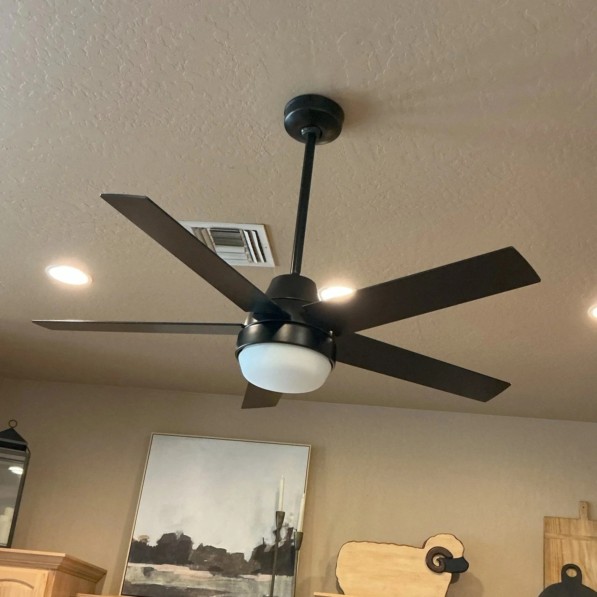 Modern black ceiling fan with a frosted glass light fixture installed on a textured beige ceiling, with recessed lighting and wooden furniture visible in the background.