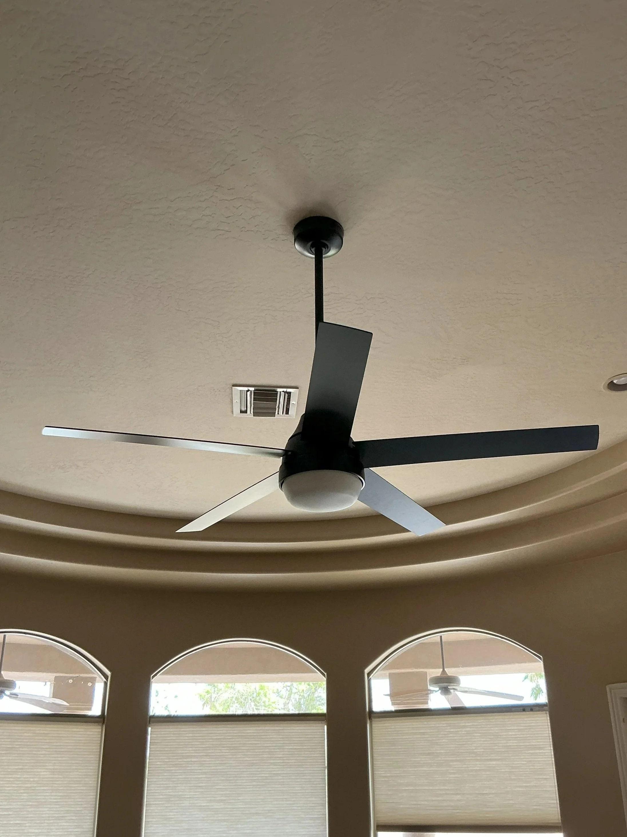 Modern ceiling fan with black and silver blades and a white light fixture, installed on a textured ceiling with windows and another fan visible in the background.