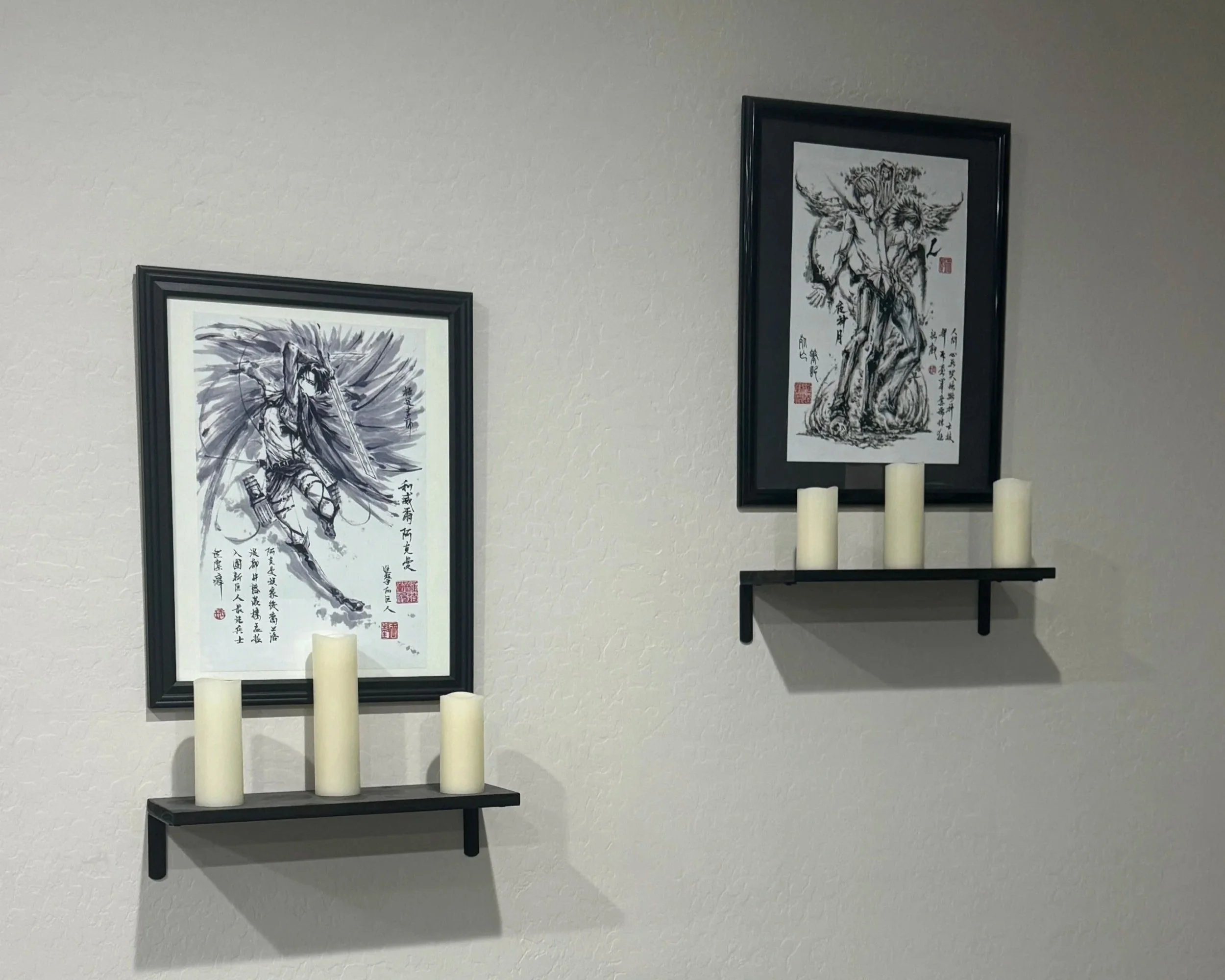 Two framed Asian-style ink paintings of mythical warriors or deities on a white wall, each with a black shelf below holding three white candles.