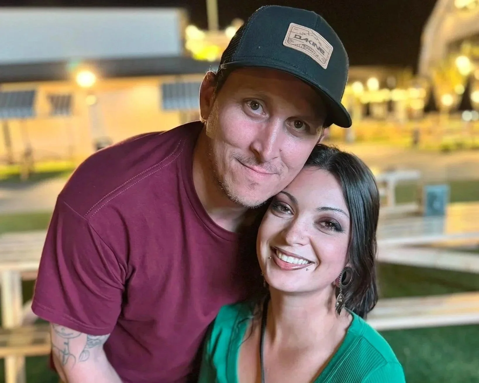A man wearing a dark green baseball cap and maroon shirt is hugging a woman with dark hair and earrings. They are smiling and outdoors at night with benches and a building in the background.