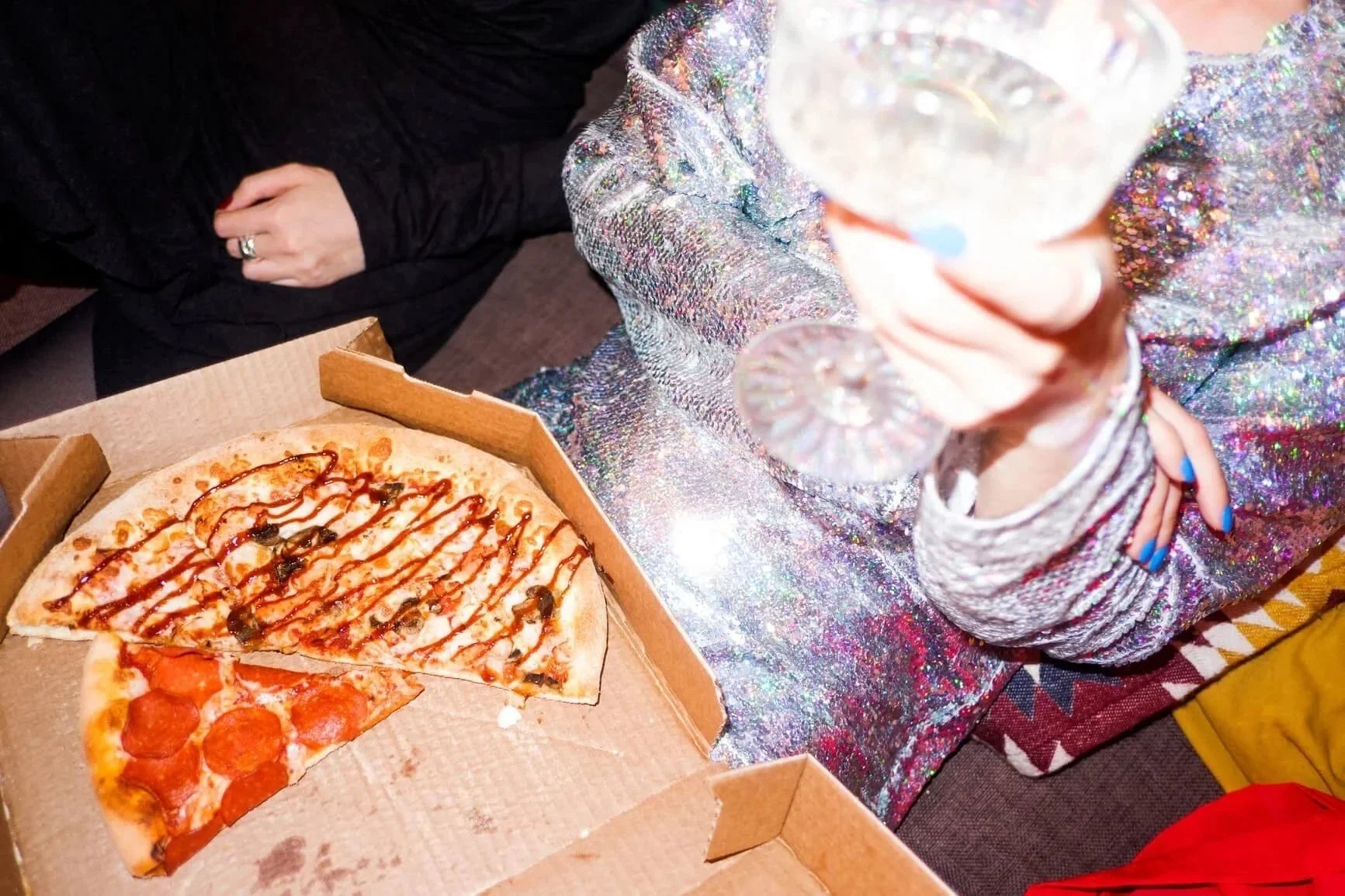 A person wearing a sparkly, colorful dress is holding a glass of beverage and sitting near a pizza in a cardboard box.