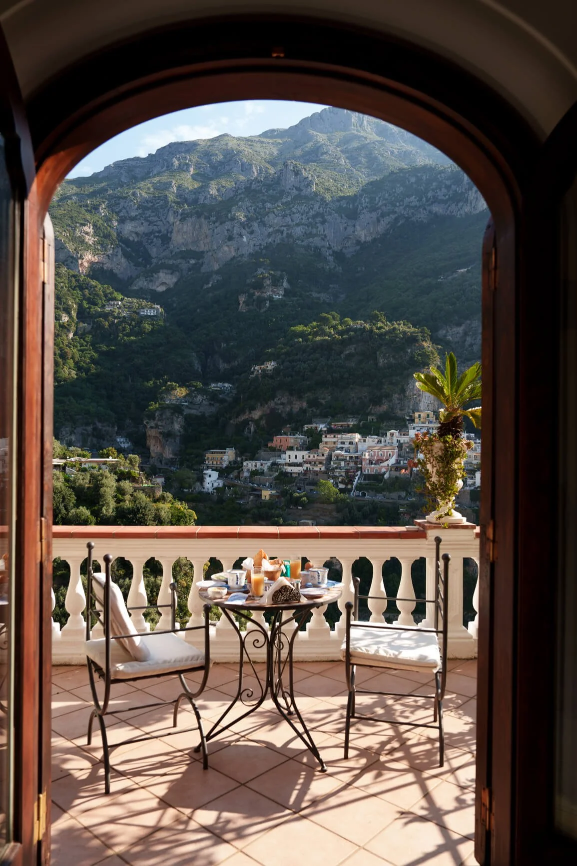A balcony with a small round table set for breakfast, overlooking a mountain village with colorful houses on a hillside, framed by an arched doorway.