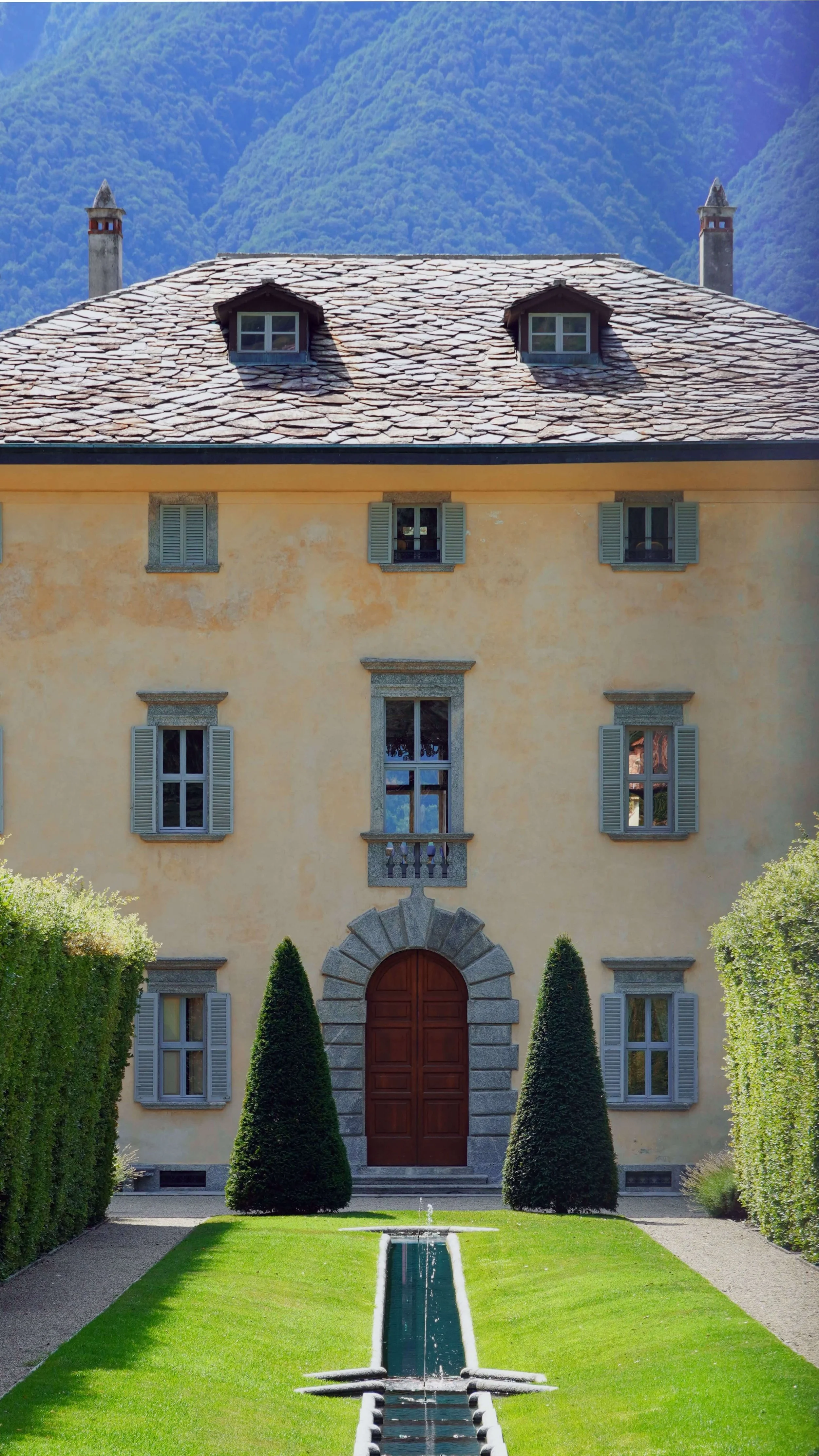 A grand yellow house with a red wooden door, surrounded by tall green bushes, with a fountain in the front yard, and mountains in the background.