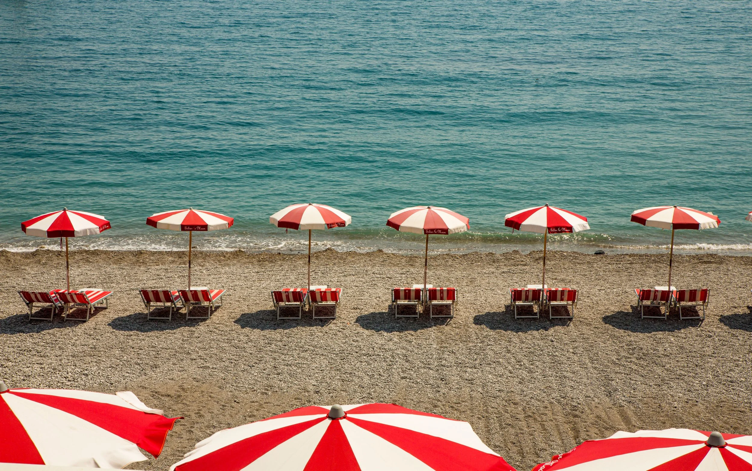 Red and white striped beach umbrellas with lounge chairs on a sandy beach near the ocean.