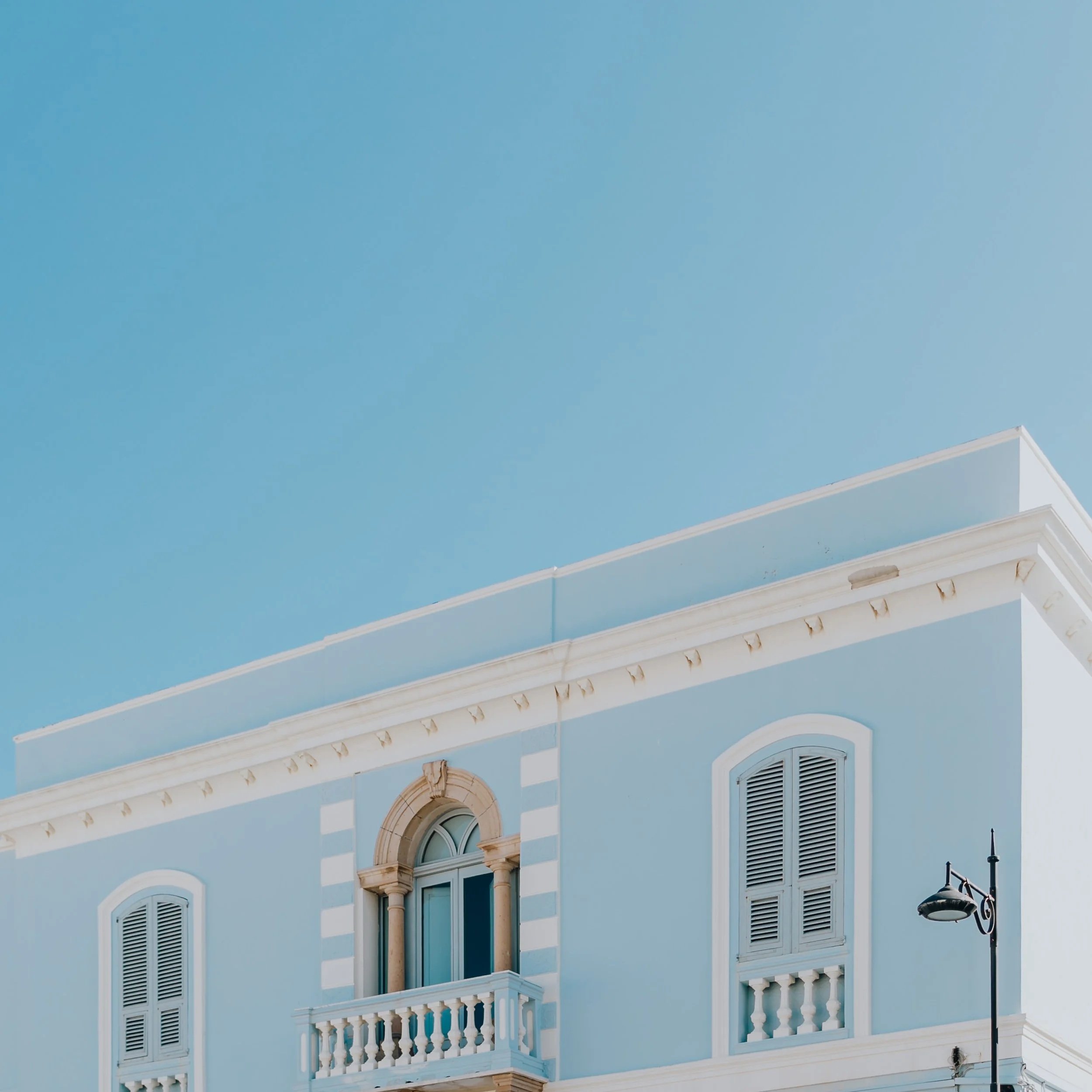 A light blue building with arched windows and white decorative trim, featuring a balcony with white balustrades and ornate architectural details under a clear blue sky.