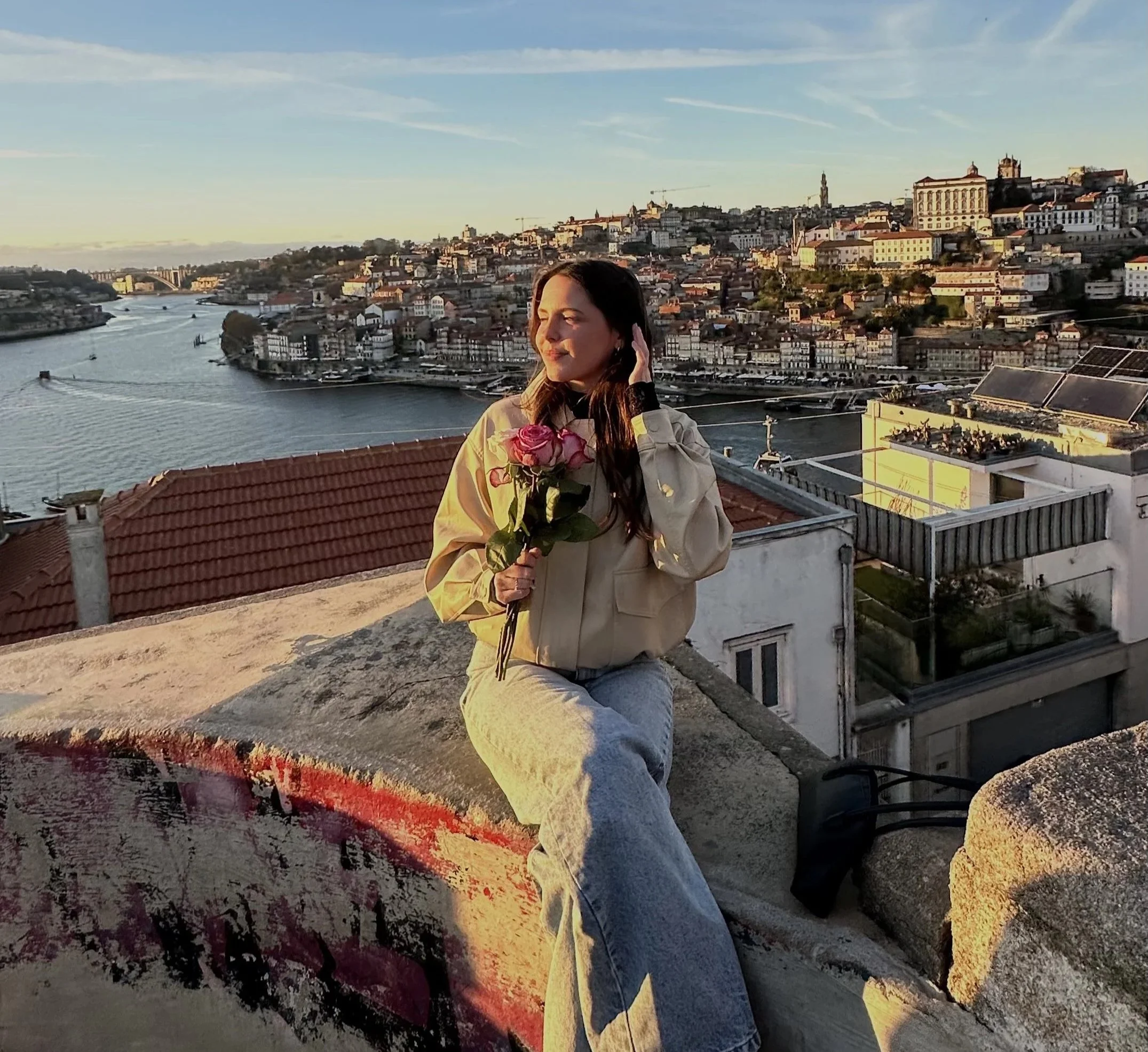 A woman sitting on a rooftop holding a bouquet of pink roses, with a cityscape and river in the background during sunset.