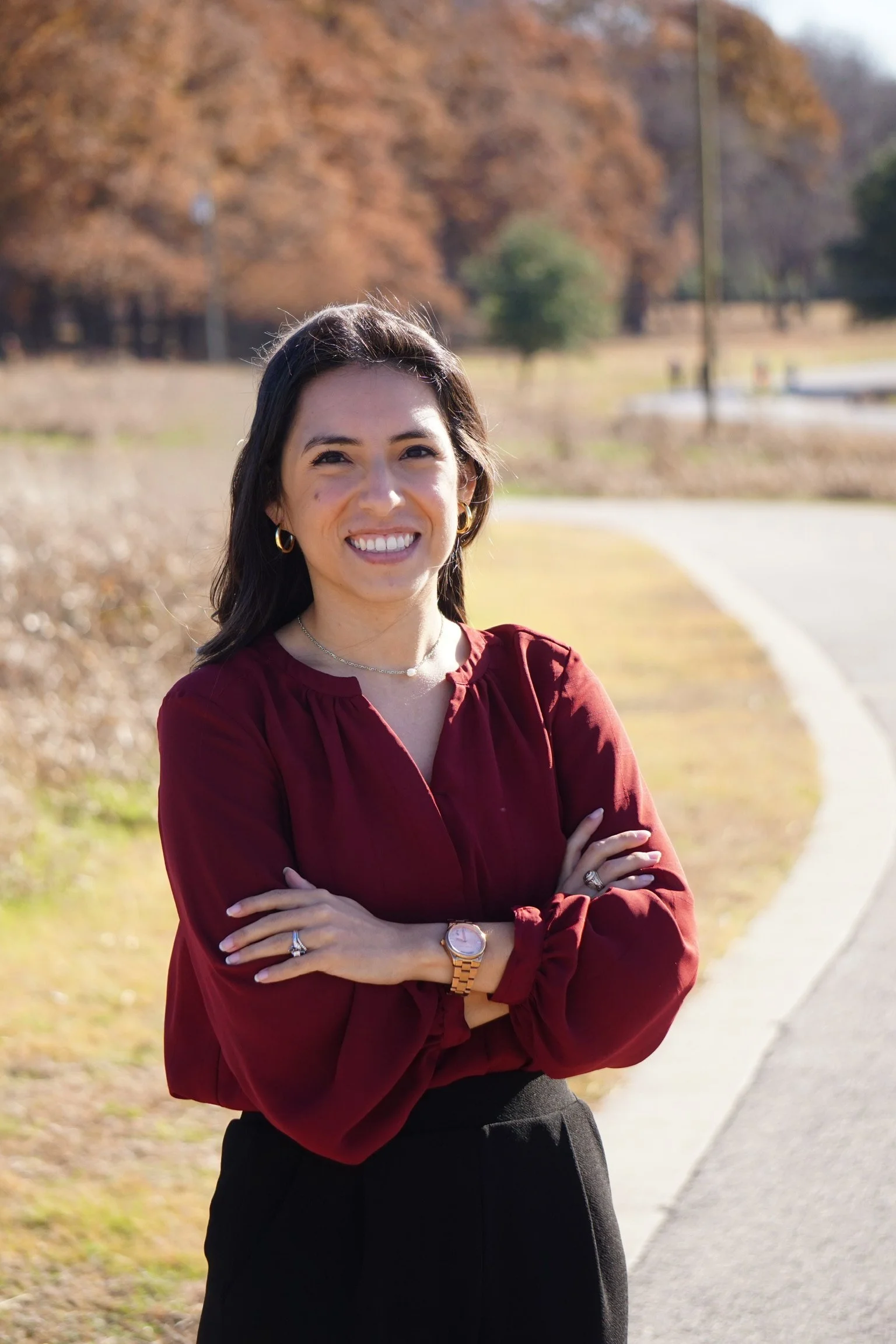 A woman with dark hair smiling outdoors in fall attire, crossing arms, with trees in autumn colors in the background.