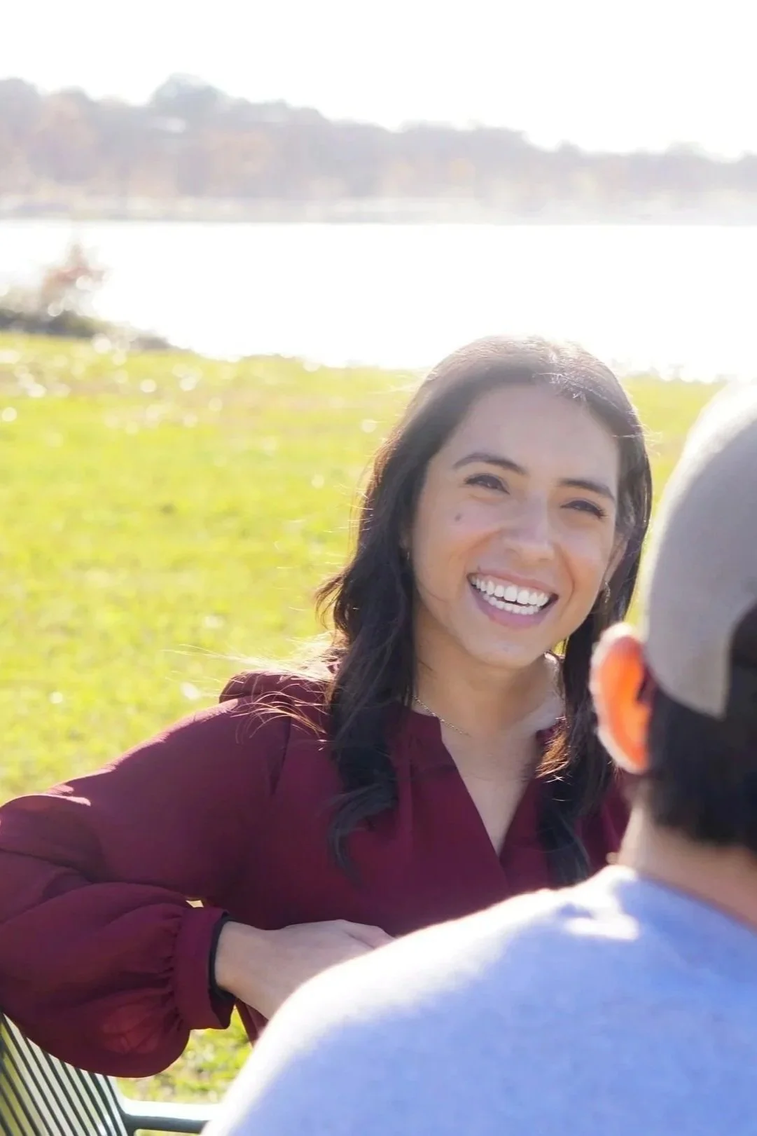 A woman with long dark hair, smiling and sitting outdoors by a body of water, wearing a maroon top, with another person partially visible in front of her.