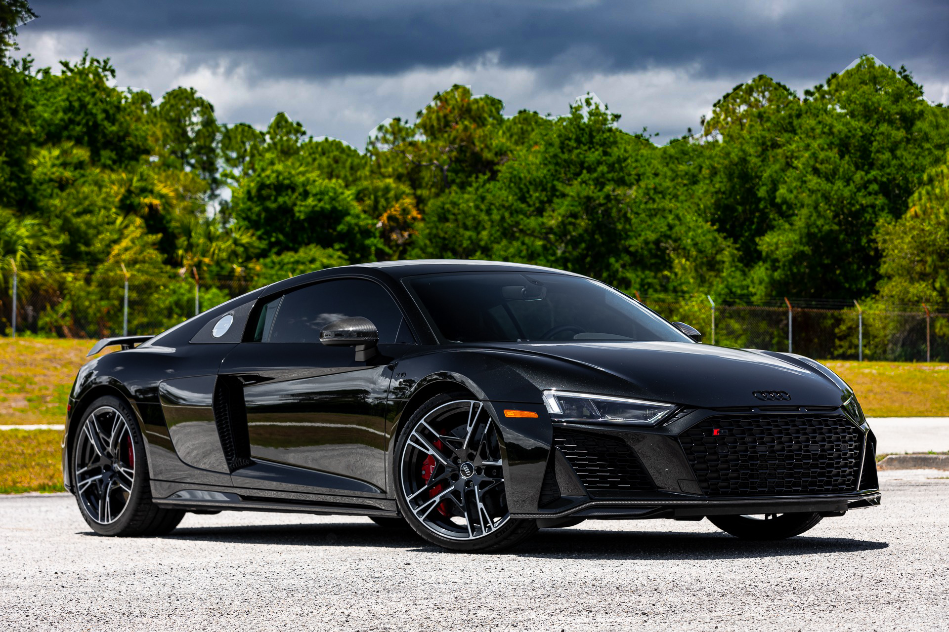Black Audi R8 parked on a paved surface with a green tree line in the background under a cloudy sky.