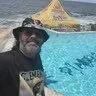 Man wearing a hat and sunglasses near a swimming pool with a yellow canopy in the background by the sea.