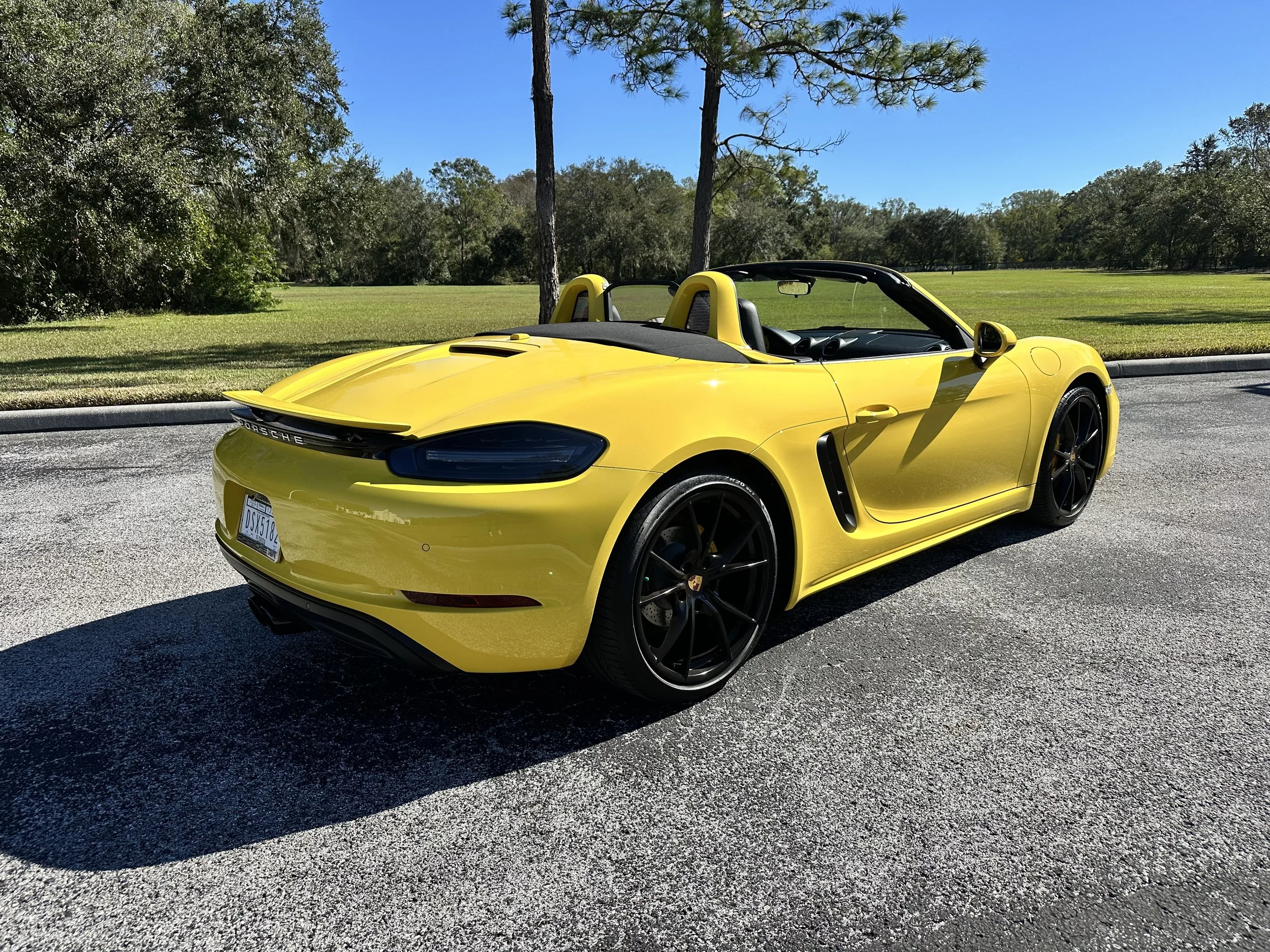 Yellow convertible sports car parked on a paved surface with a grassy field and trees in the background.