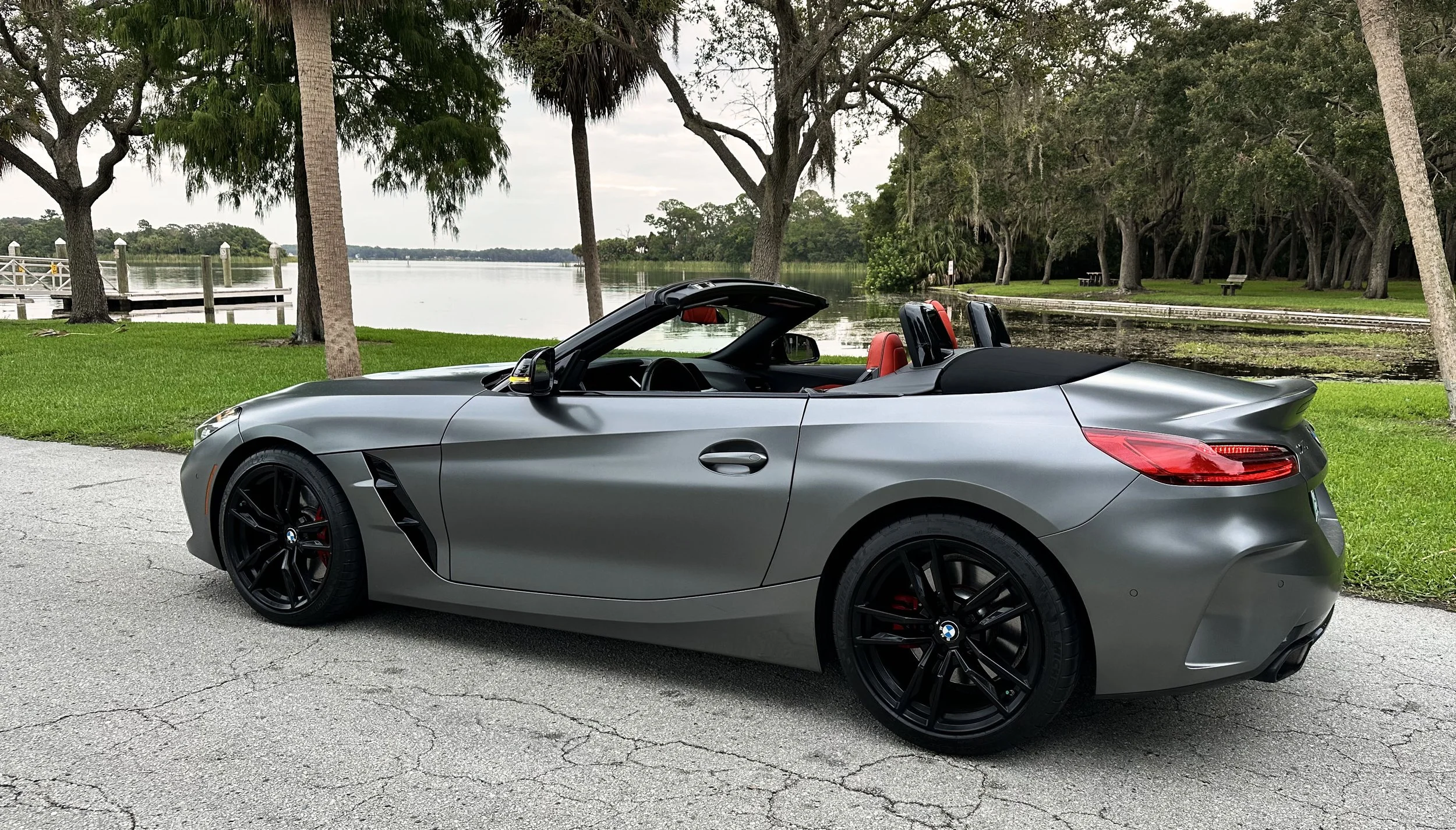 A silver convertible car parked near a lake with trees in the background.