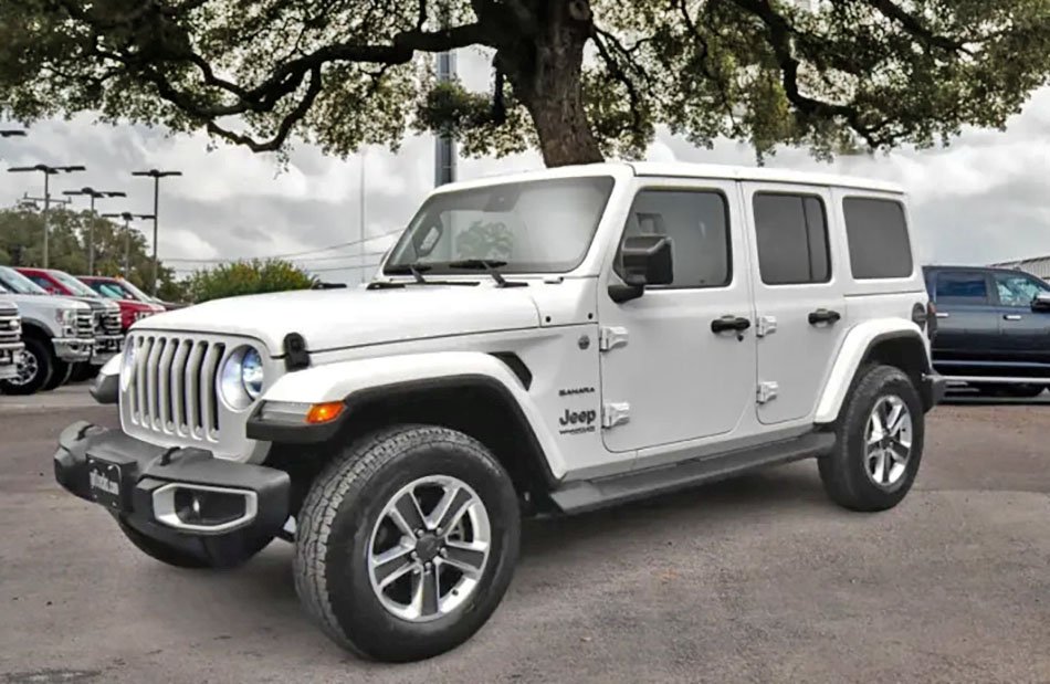 White Jeep Wrangler SUV parked under a tree next to other vehicles at a dealership lot.