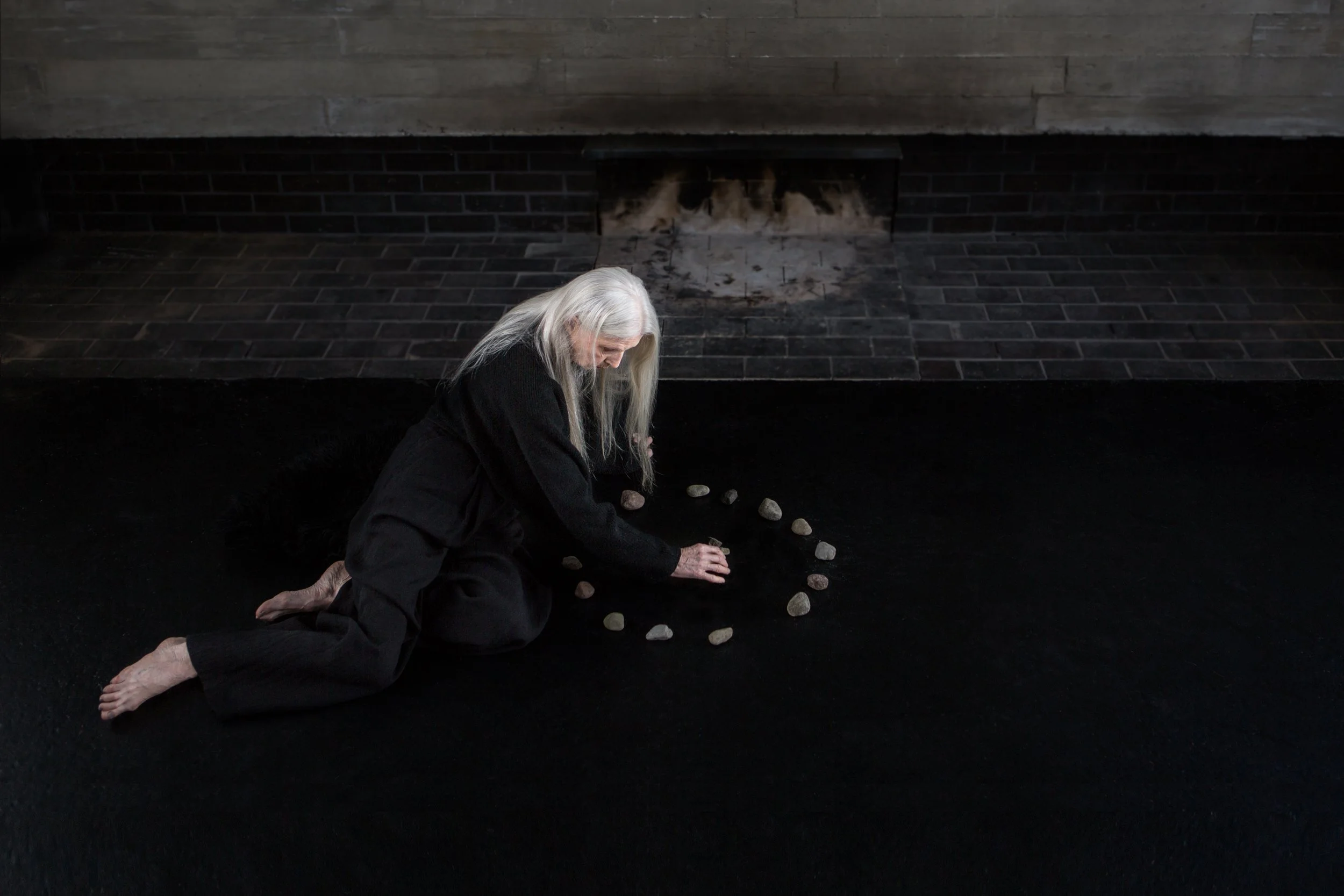 An elderly woman with long white hair kneels on the floor, arranging a line of small rocks in front of her. She is barefoot and dressed in black clothing. The background features a dark brick wall and a fireplace with a wooden mantle.
