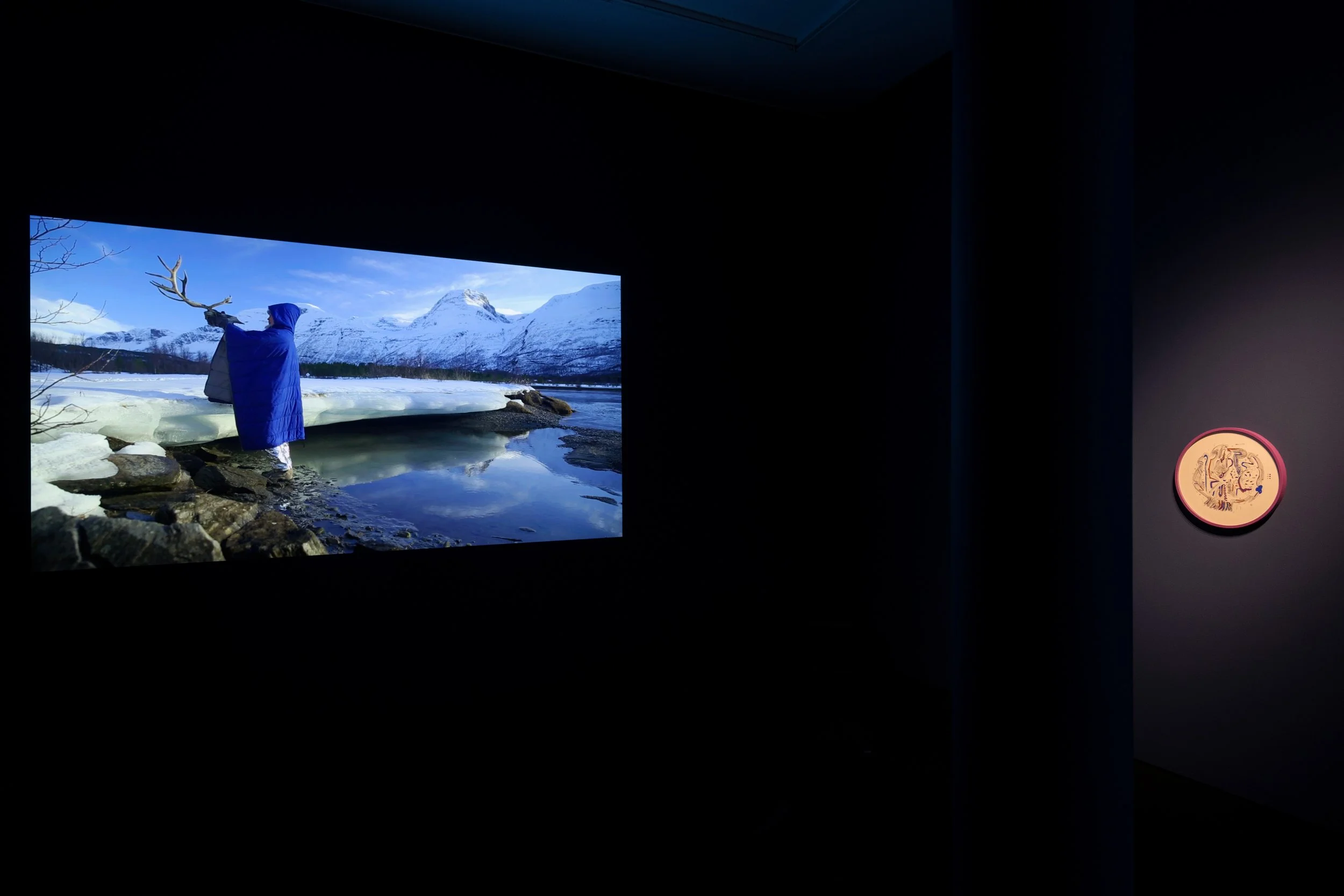 An indoor display showing a landscape photograph of snowy mountains, a river, and a person dressed in a blue coat holding a reindeer antler, framed by a black border with a coin or medallion on the right side.