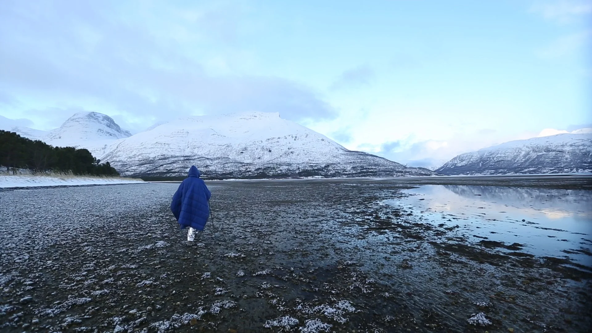 Person in a blue jacket walking along a rocky shoreline with snow-capped mountains and a calm reflective lake in the background.