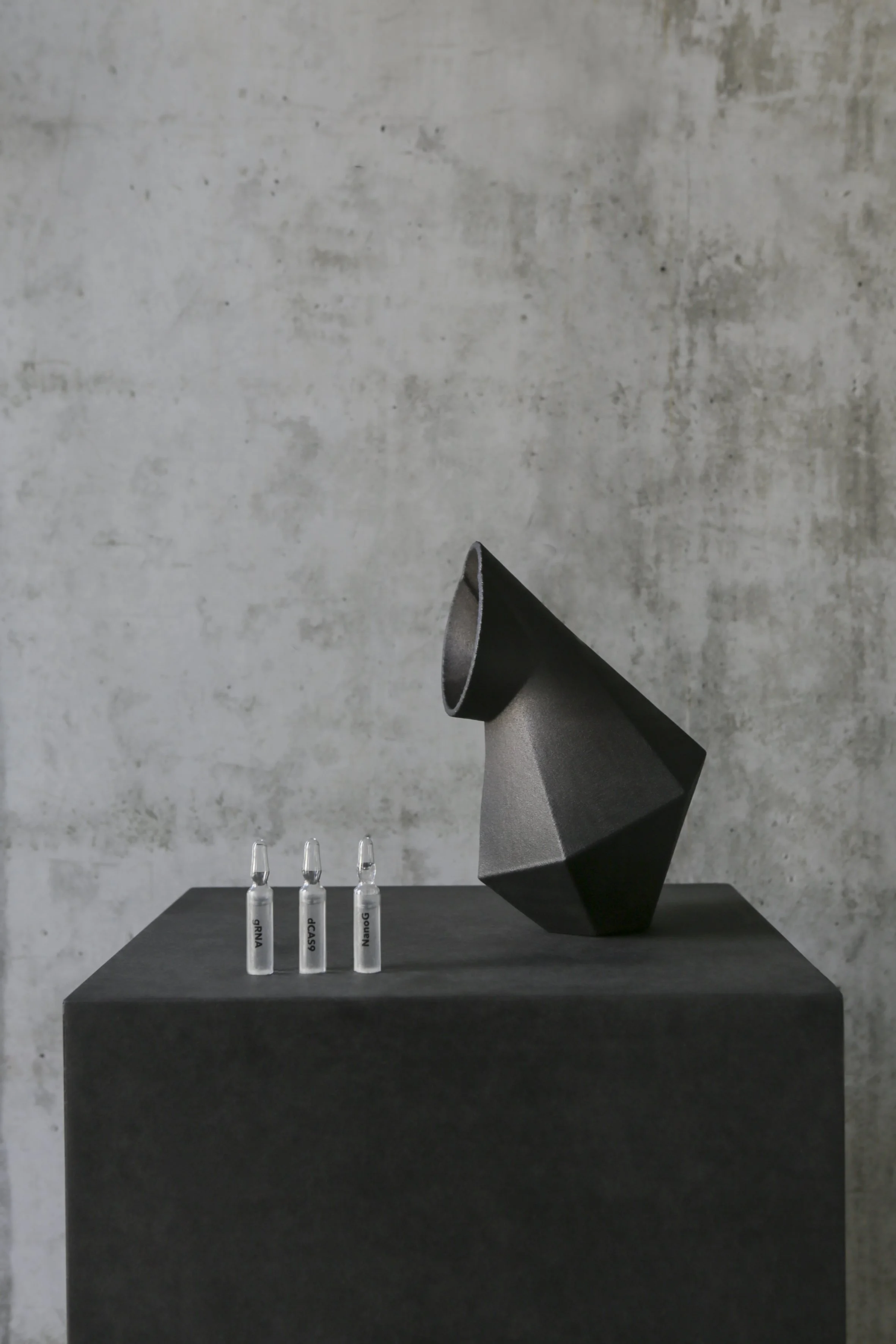 A large black sculpture shaped like a cone with a pointed end, placed on a black pedestal against a plain gray wall. In front of the sculpture, there are three small clear glass ampules with labels, standing upright on the pedestal.