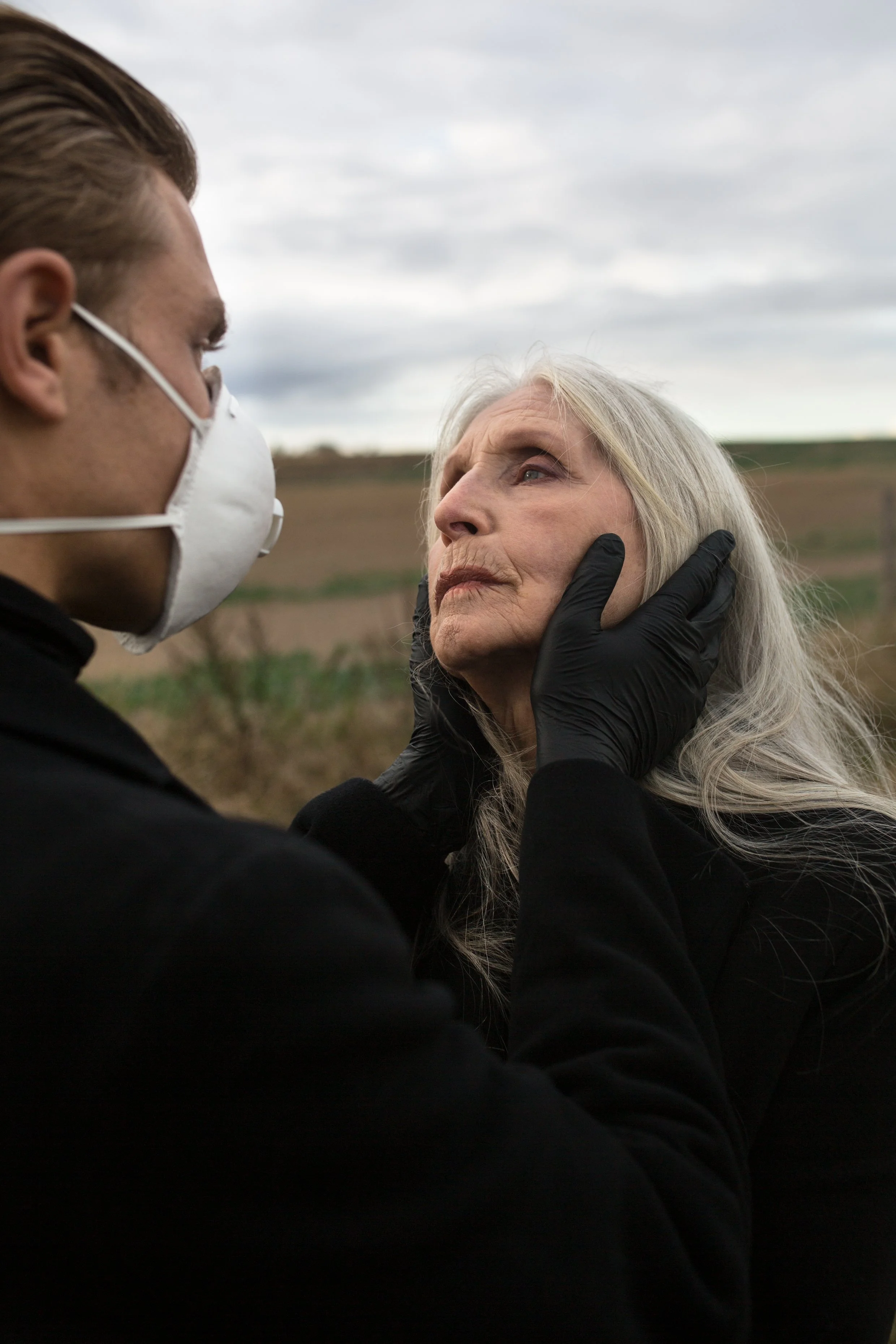 A man wearing a white face mask and black gloves gently holds and looks at an elderly woman's face outdoors, with a cloudy sky and open field in the background.