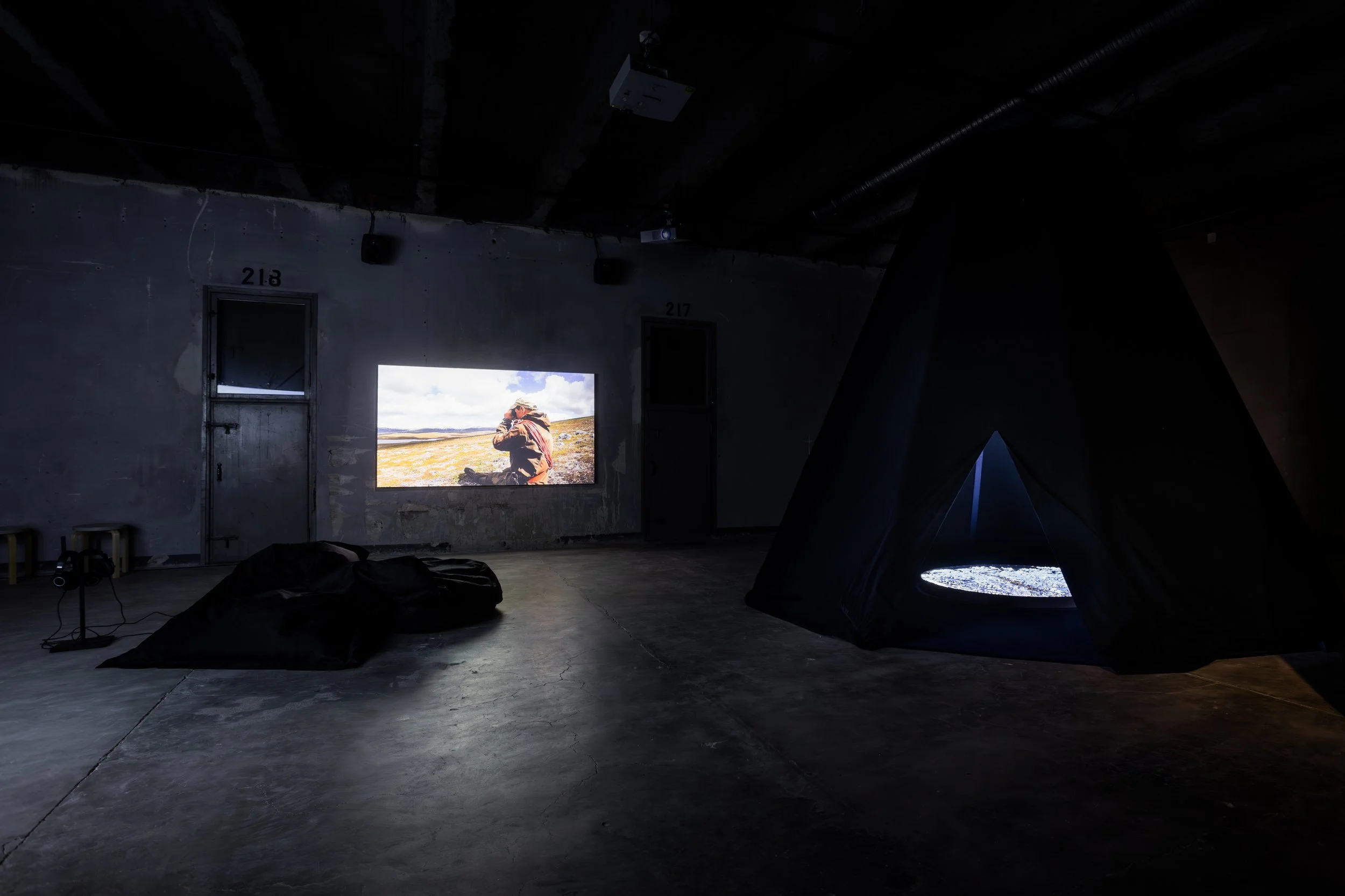 Dark room with a projection of a landscape on the wall showing a person in outdoor gear, outdoors with cloudy sky, tent on the floor, and some equipment.