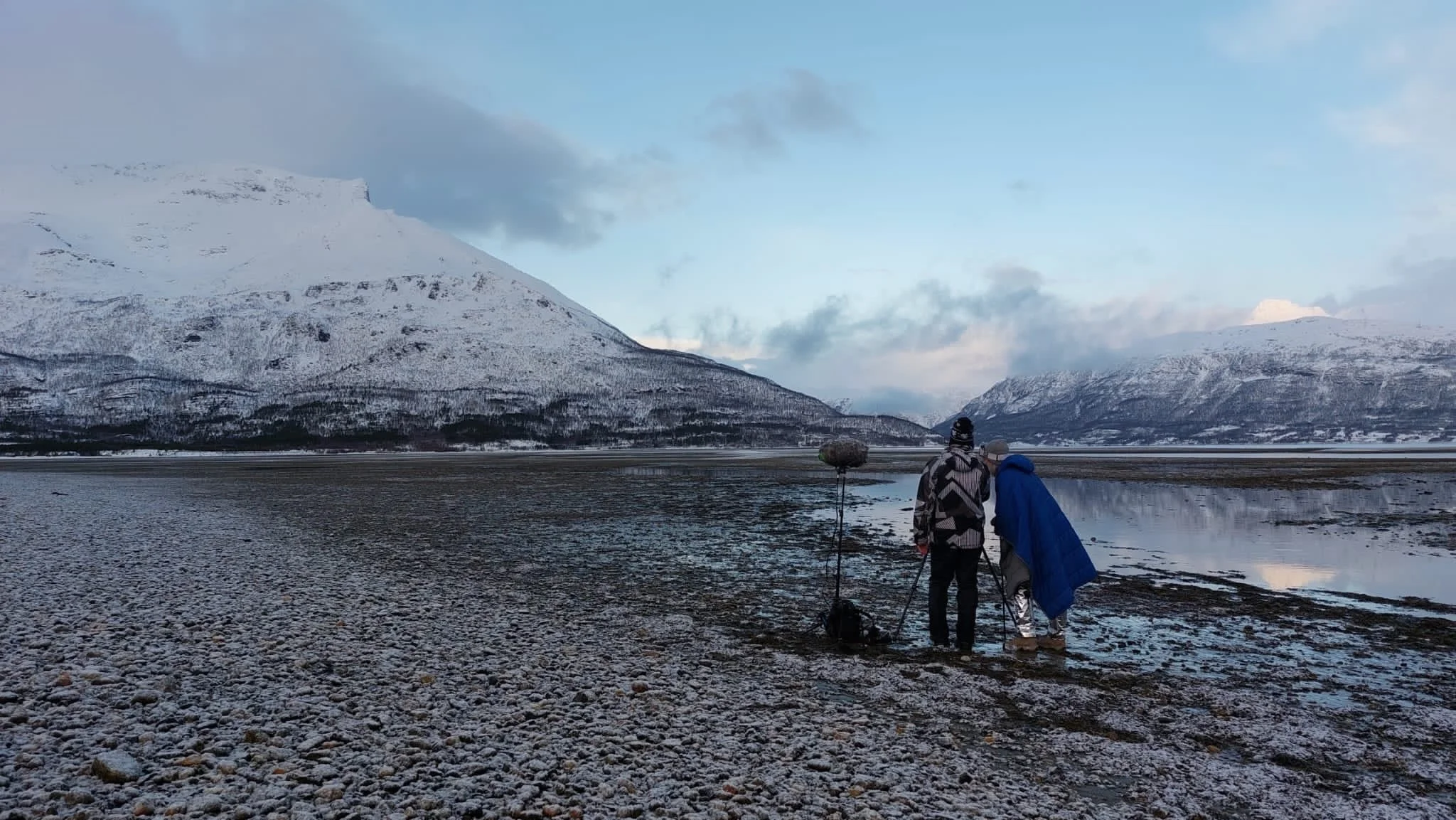 Two people with backpack and tripod set up on rocky shore by water, snow-covered mountains in the background, cloudy sky.