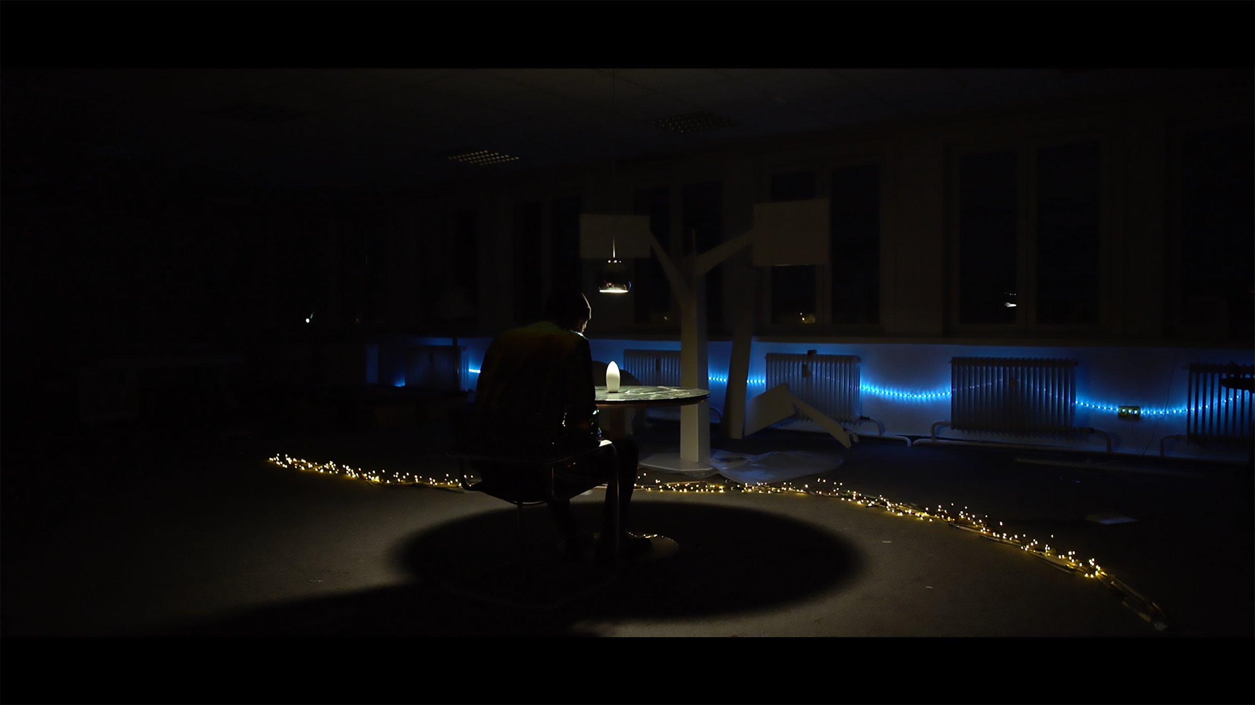A person sitting alone at a table with a lamp in a dark room decorated with blue LED string lights and small yellow fairy lights on the floor, with some furniture and radiators visible.