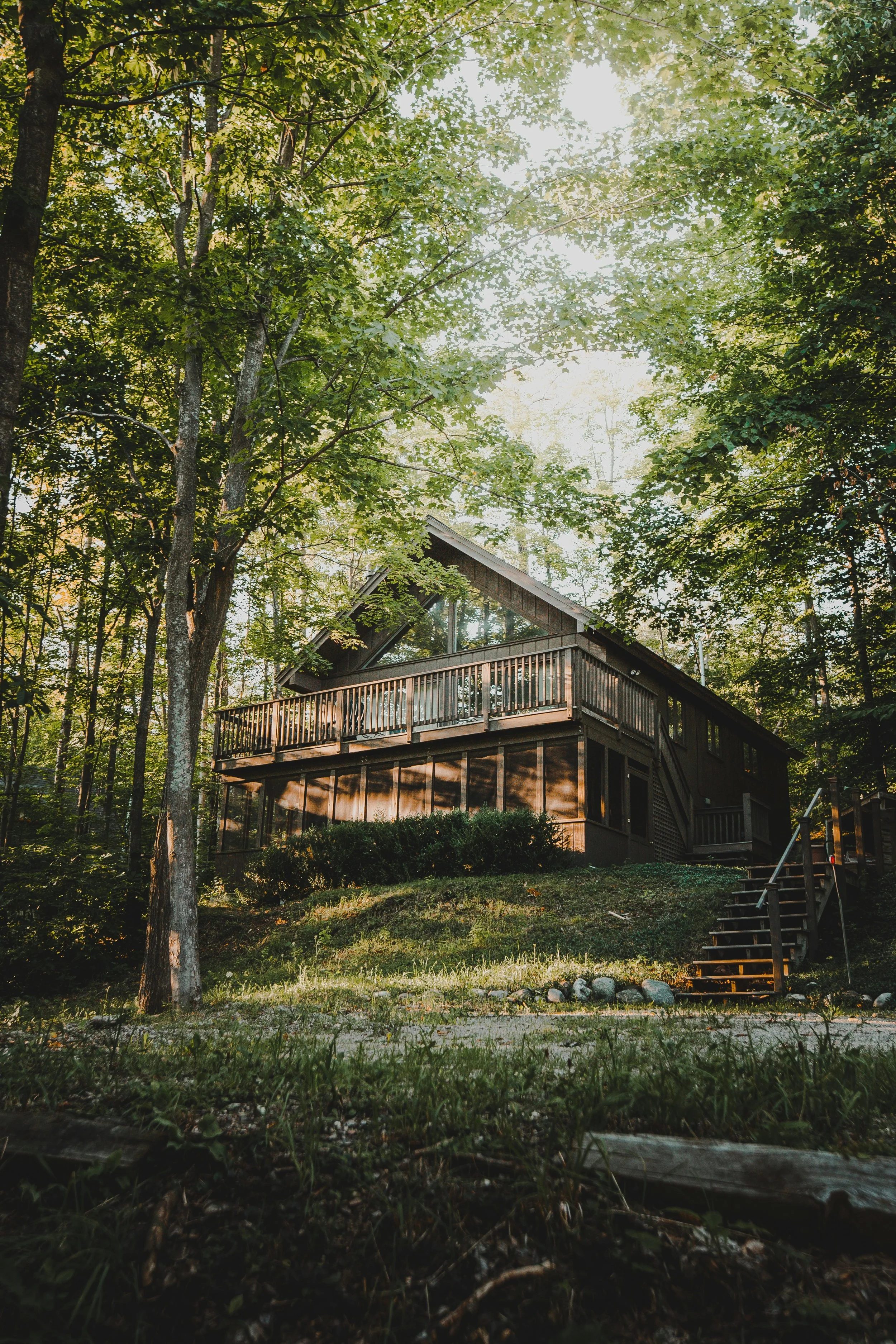 A wooden house with a large balcony is situated among trees in a forest, with sunlight filtering through the leaves.