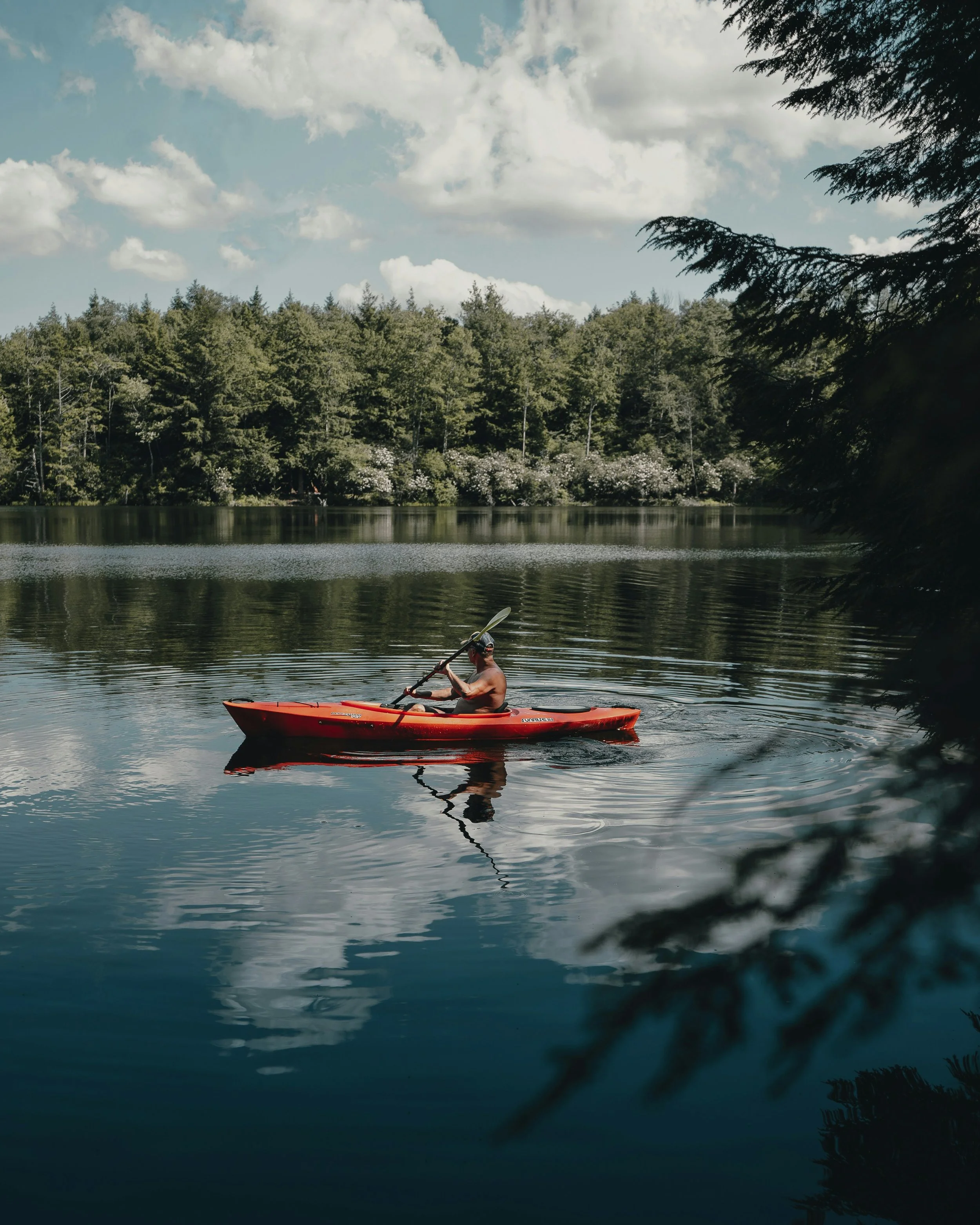 A person kayaking on a calm lake with a forest and cloudy sky in the background.